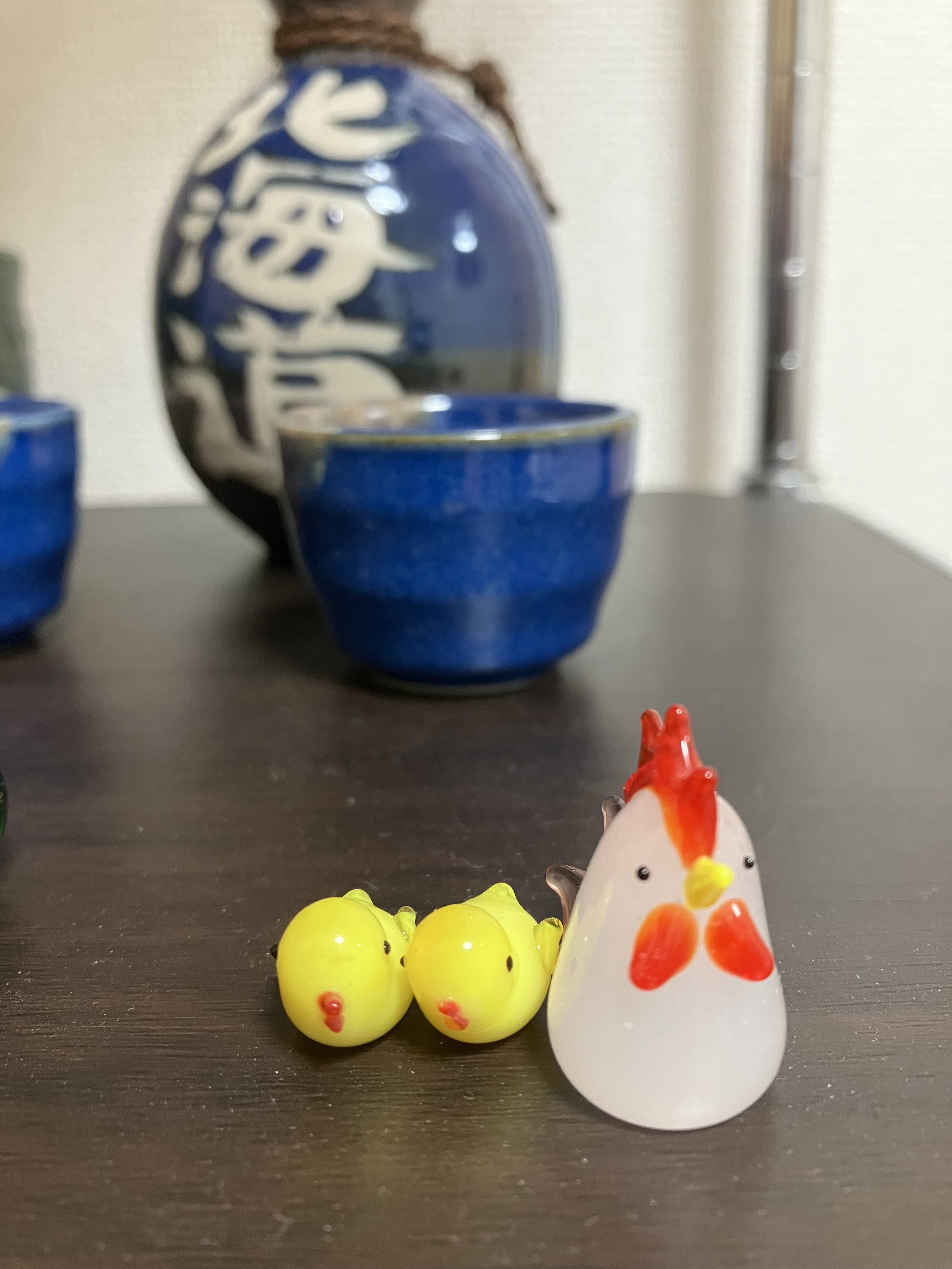 Small decorative glass figures of a chicken and three chicks on a dark wooden surface with blue ceramic bowls and a blue decorative vase in the background.