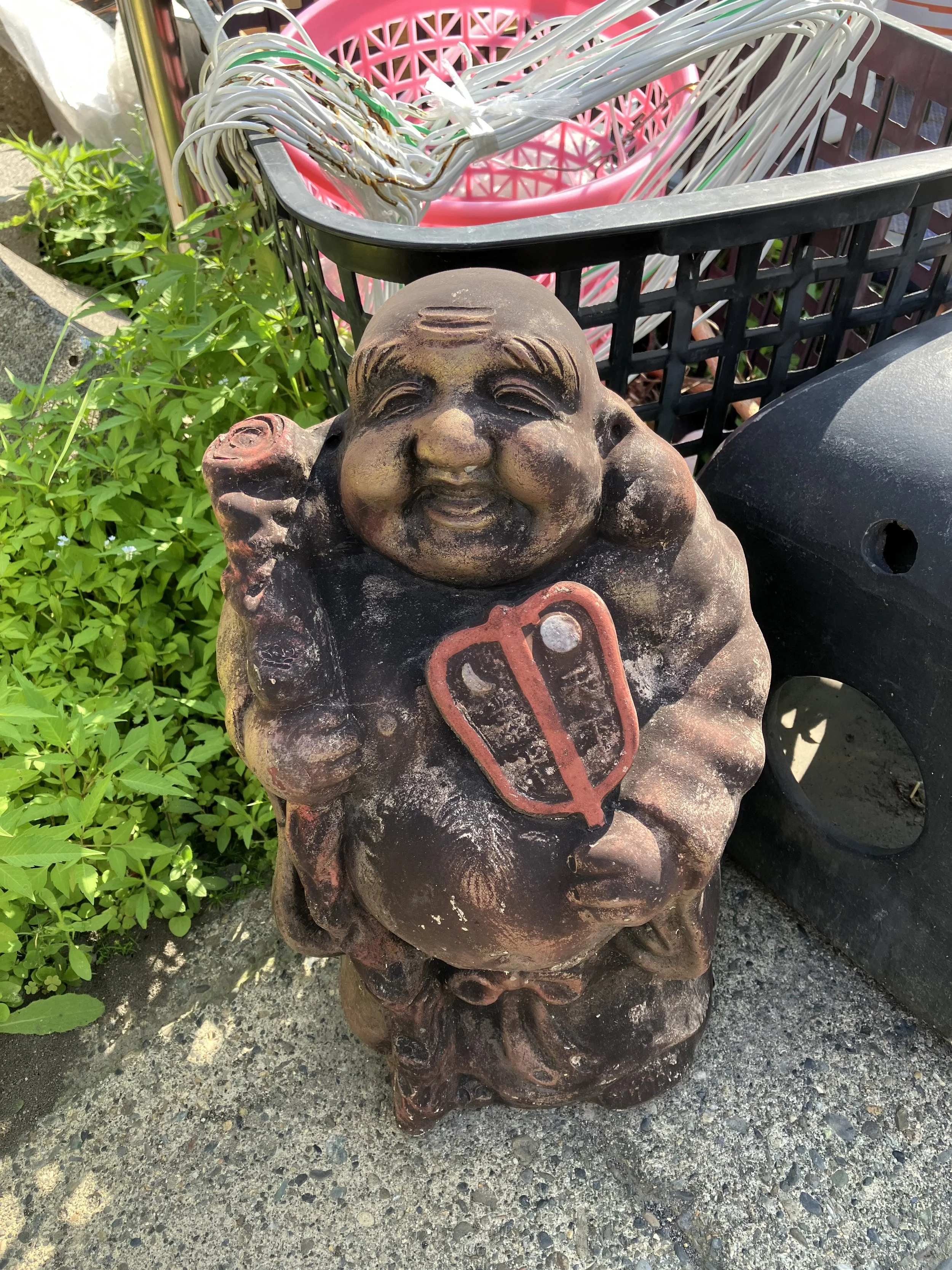 A weathered statue of a smiling, chubby, cartoon-like boy with a large head, holding what appears to be a club in one hand and a shield with the letter 'Q' on it in the other, placed outdoors near greenery and shopping carts.
