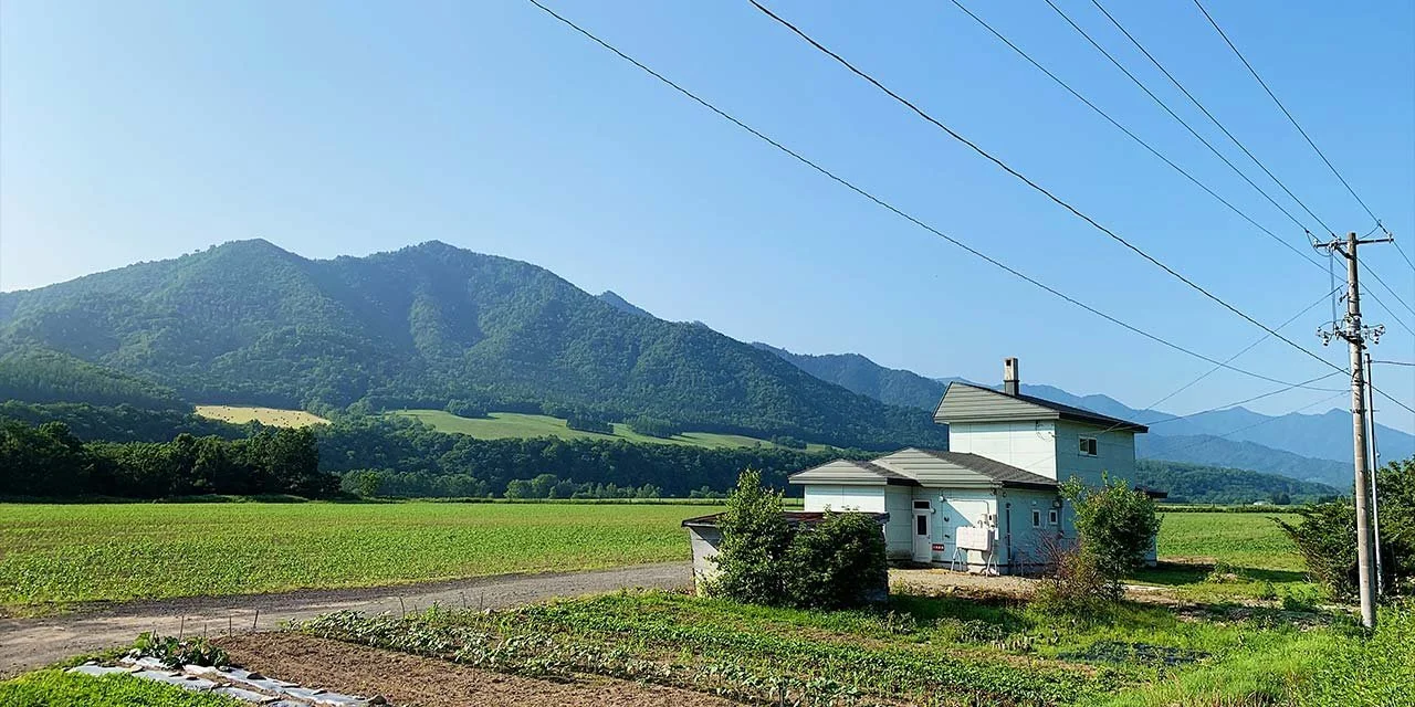 A rural landscape with a white house, green fields, and mountains in the background under a clear blue sky.