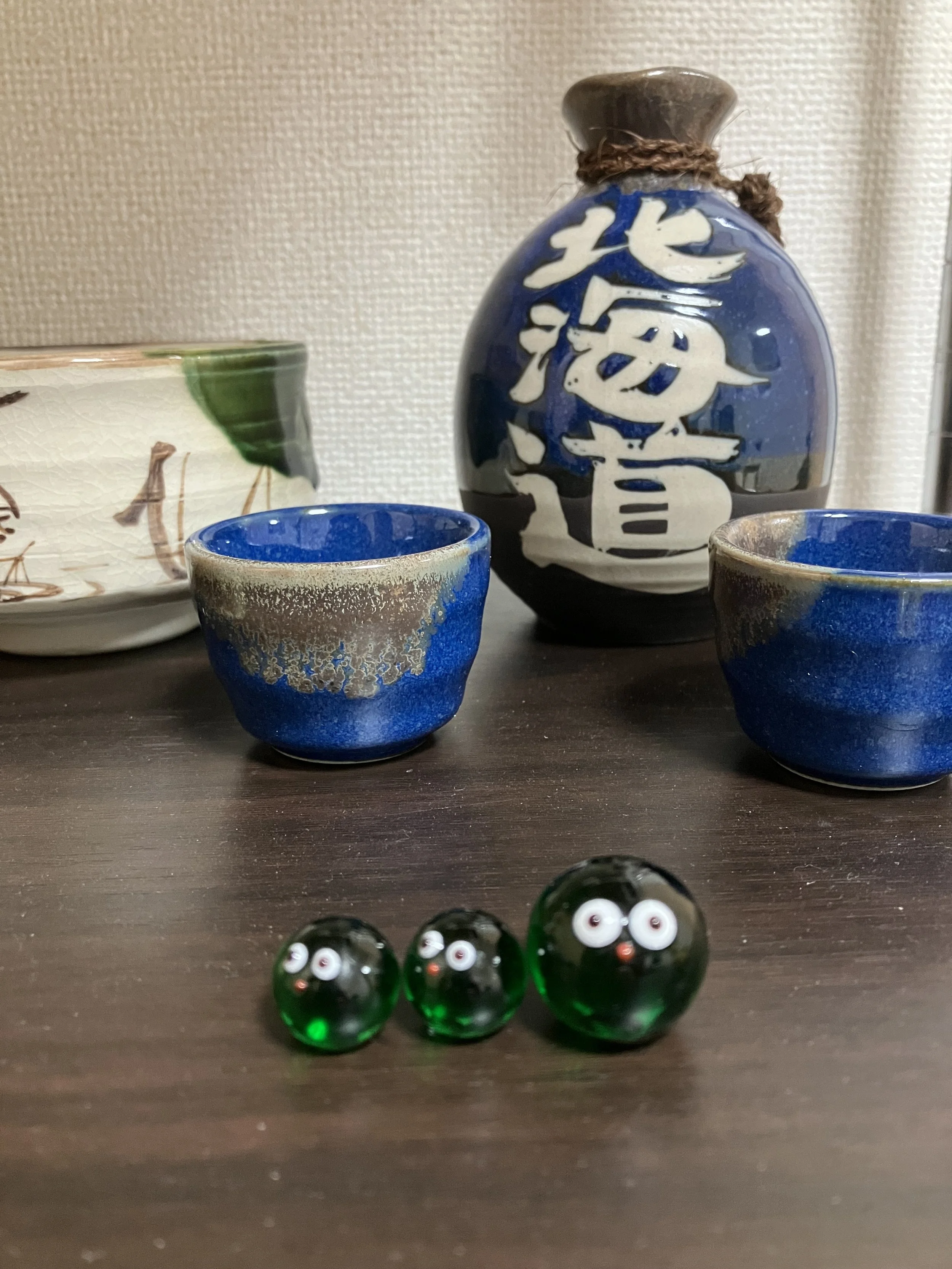 Decorative ceramic and glass items including a blue and white bottle with Chinese characters, two small blue ceramic cups, a partially visible white bowl with green accents, and three small green glass balls with googly eyes on a wooden surface.