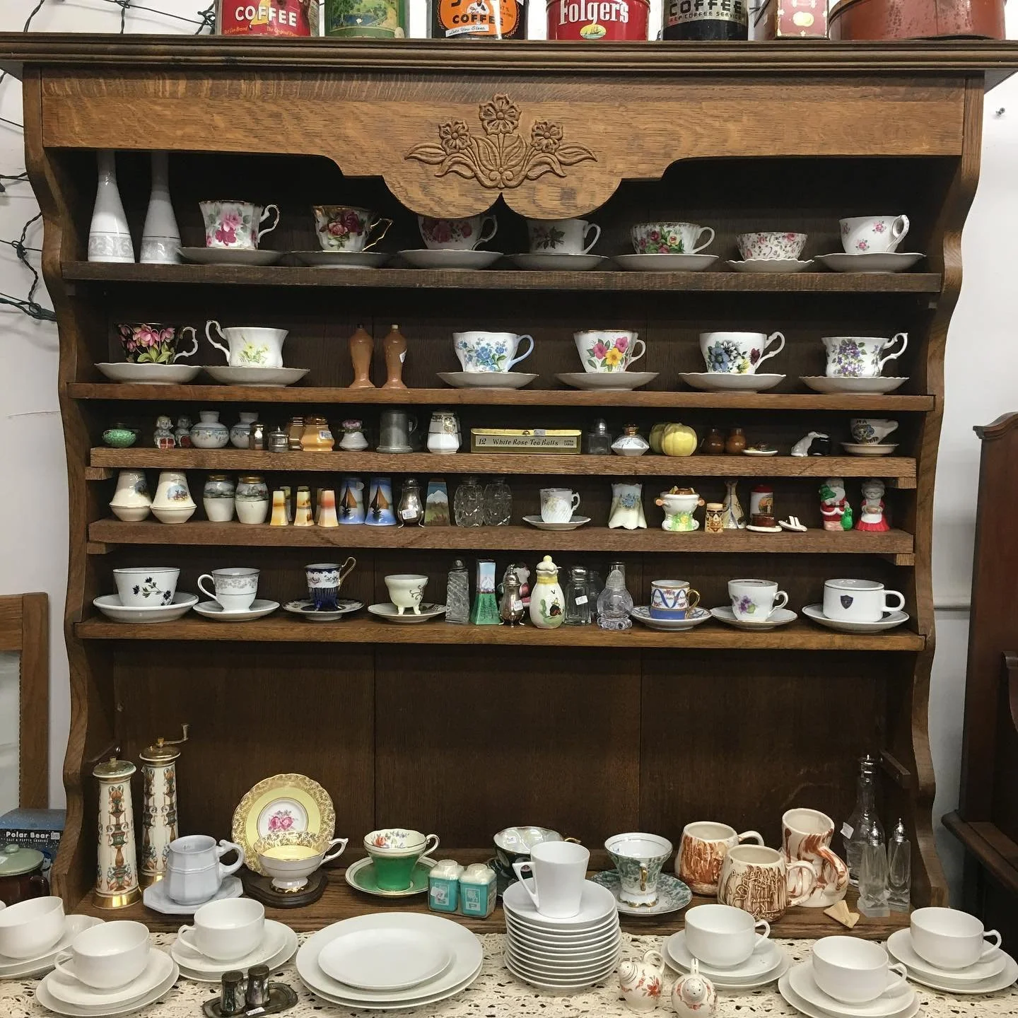 A wooden shelf filled with various decorative teacups, teapots, and small collectibles, with larger dishes and cups arranged on a lace tablecloth in front.