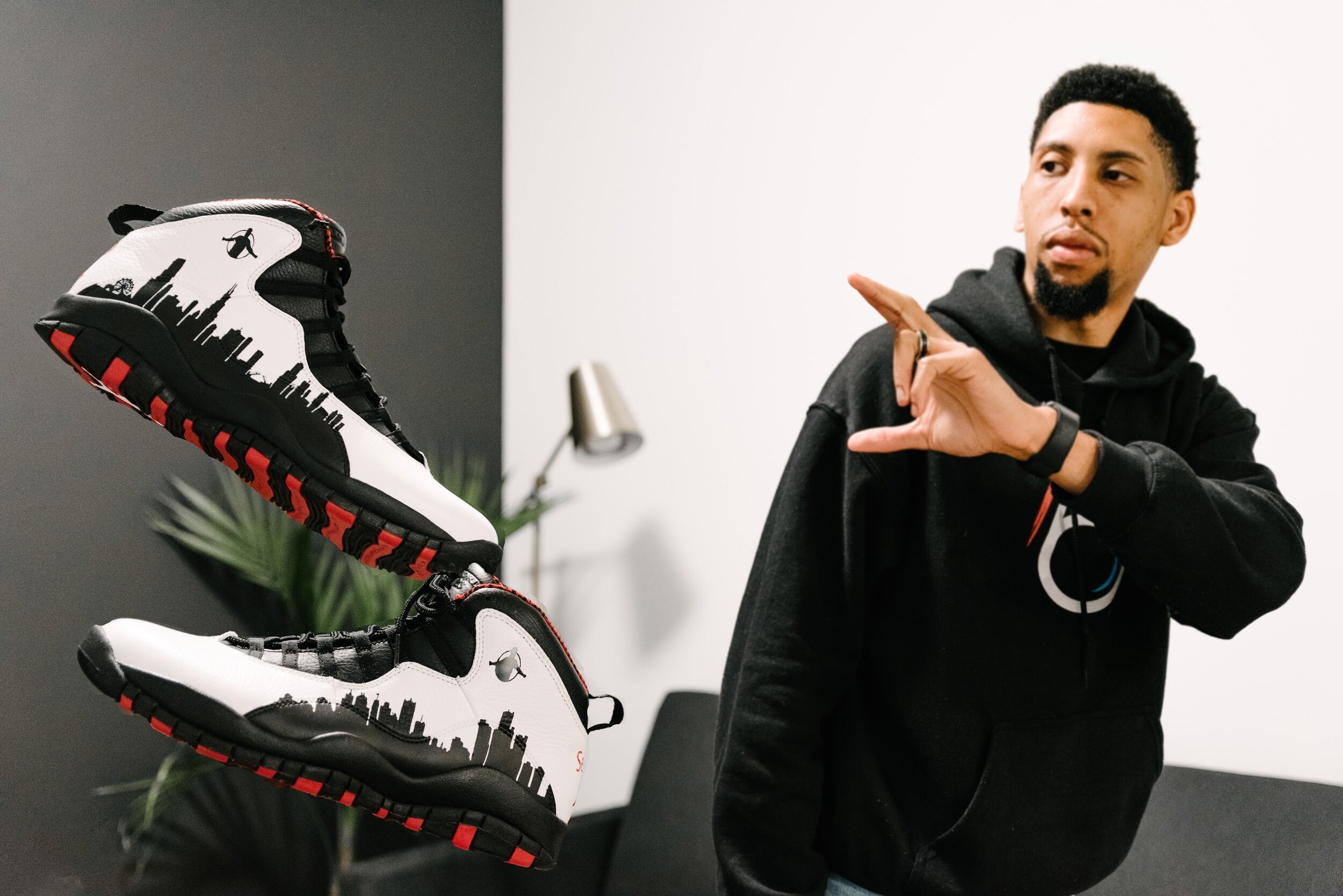 A man wearing a black hoodie gestures with his right hand, while two Nike sneakers with city skyline and bee logos hover in front of him indoors.