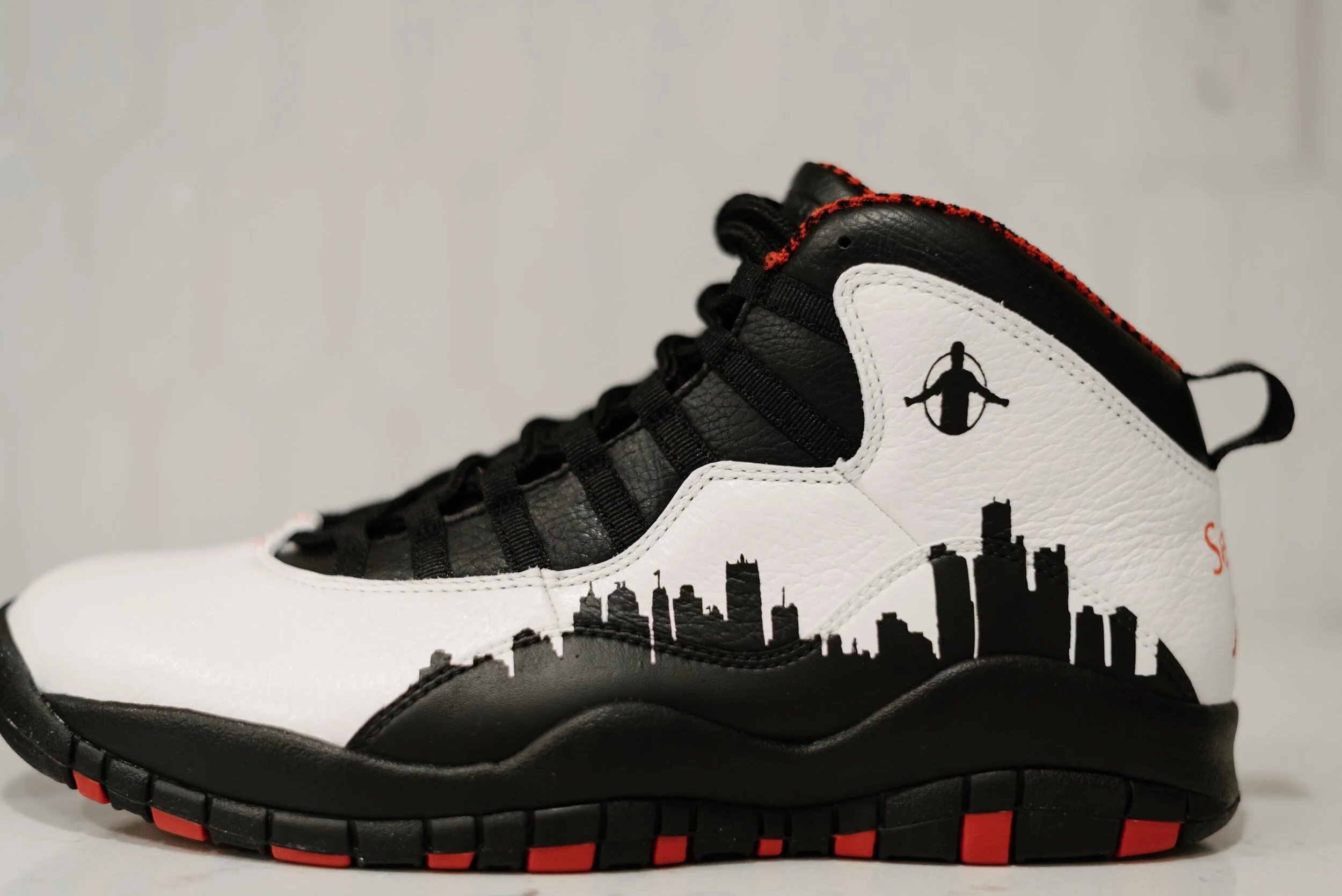 Black and white sneaker with city skyline and skyline silhouette on side, featuring black laces and red accents on sole, high-top design with pull tab at back.