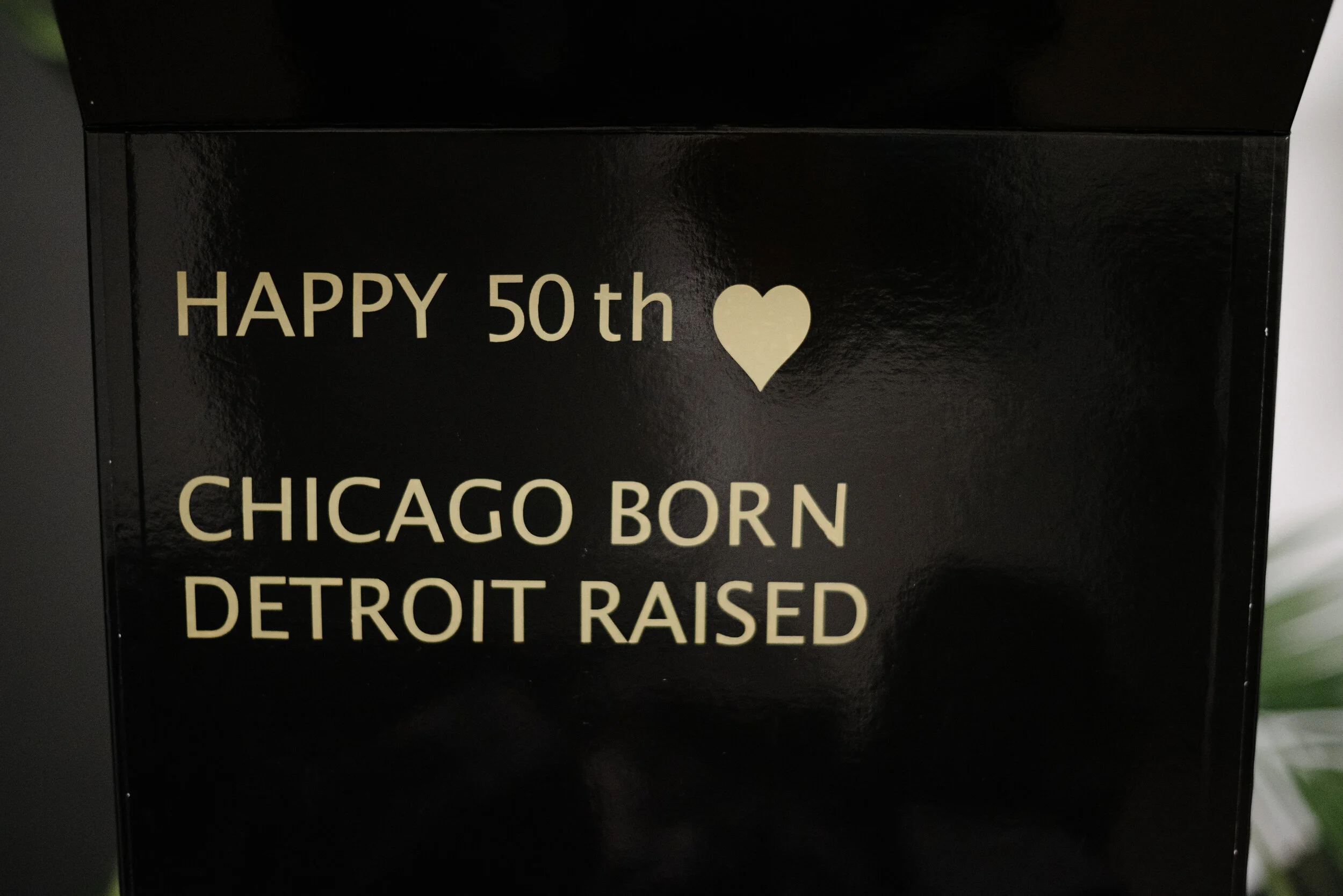 A black box with gold-colored text that reads: 'HAPPY 50th ❤️ CHICAGO BORN DETROIT RAISED'