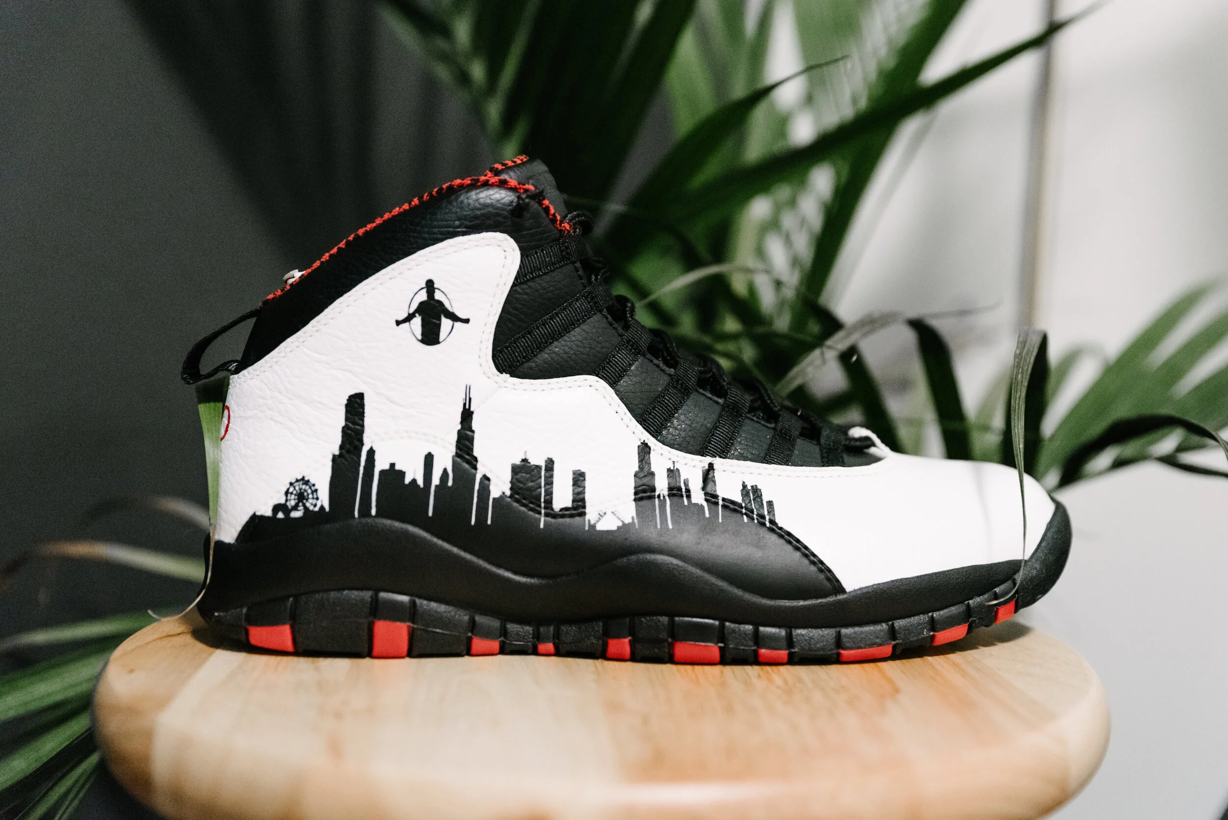 Black and white athletic shoe with a city skyline design and a small bee logo, placed on a wooden surface with green plants in the background.