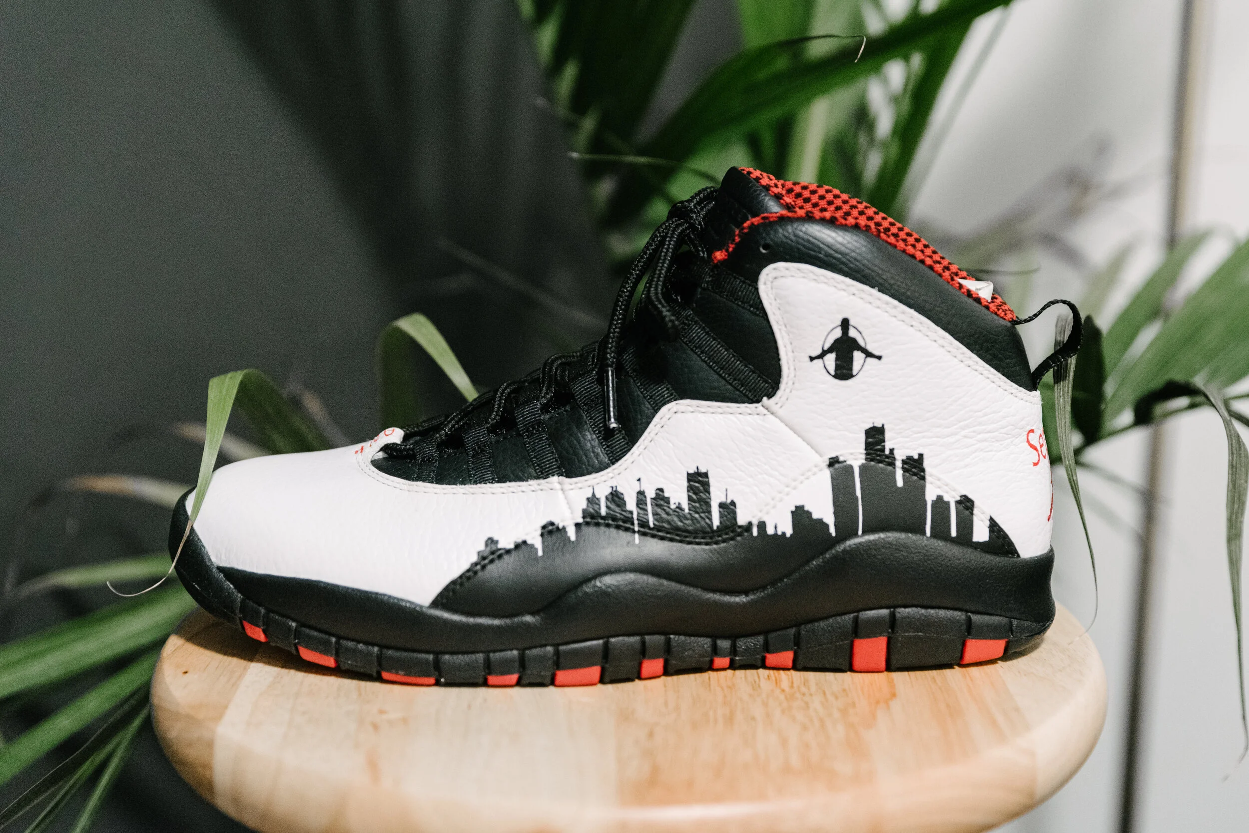 A sneaker with a city skyline design in black and white on the side, placed on a wooden stool with green plants in the background.
