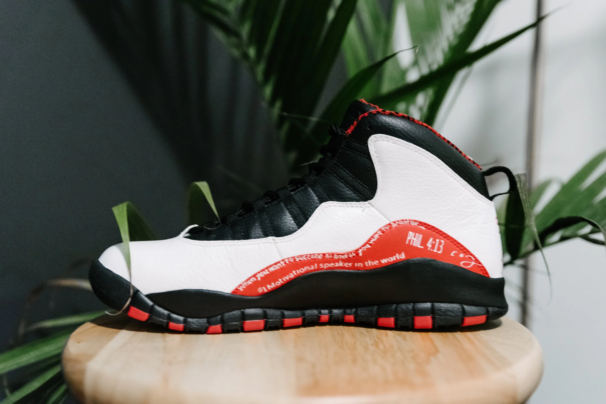 A black, white, and red athletic shoe with motivational text on the side, placed on a wooden surface with green plants in the background.