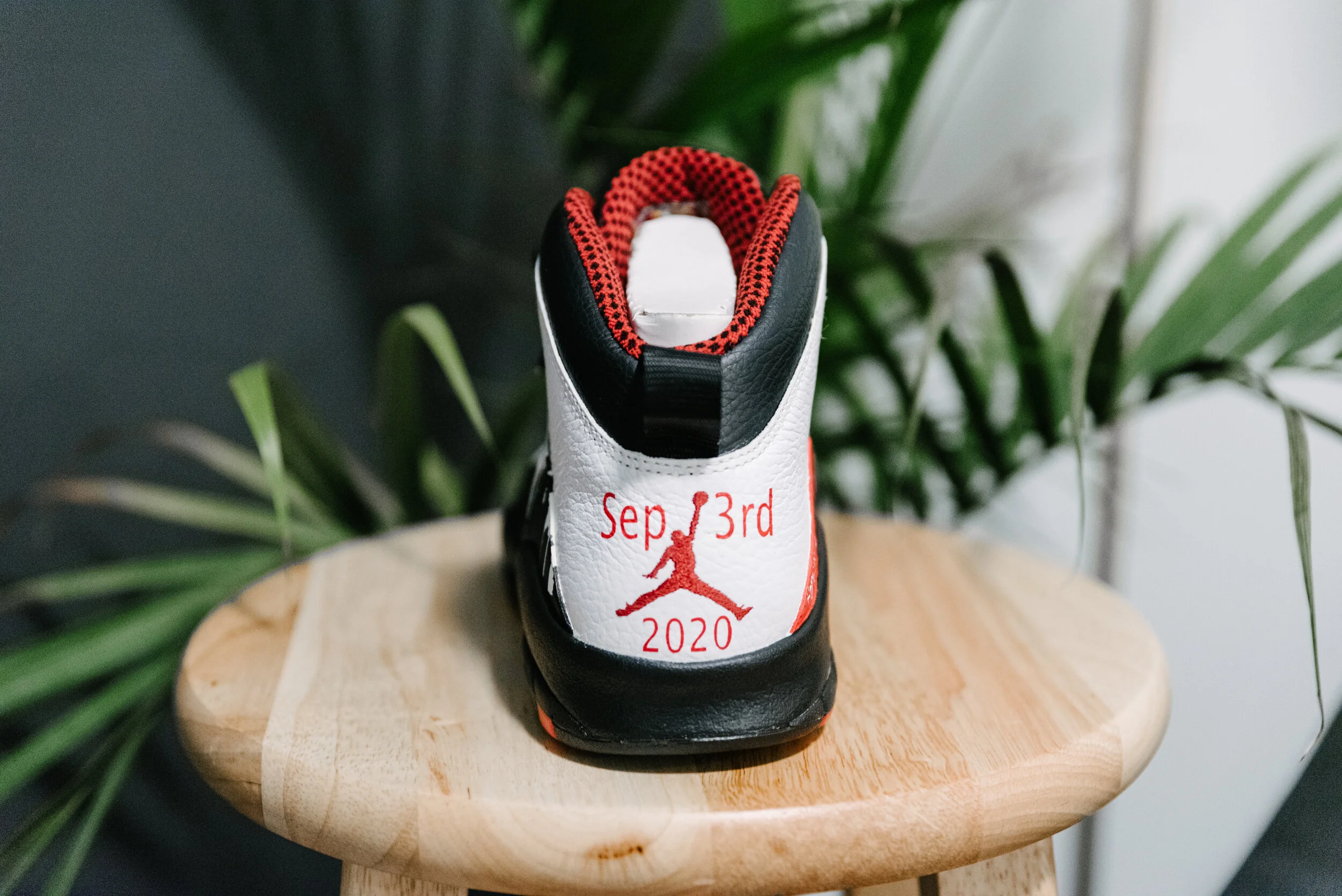 Back view of a custom sneaker with a embroidered date 'Sep 3rd 2020', a silhouette of a person jumping, and details of red, white, and black colors, placed on a wooden stool with green plants in the background.