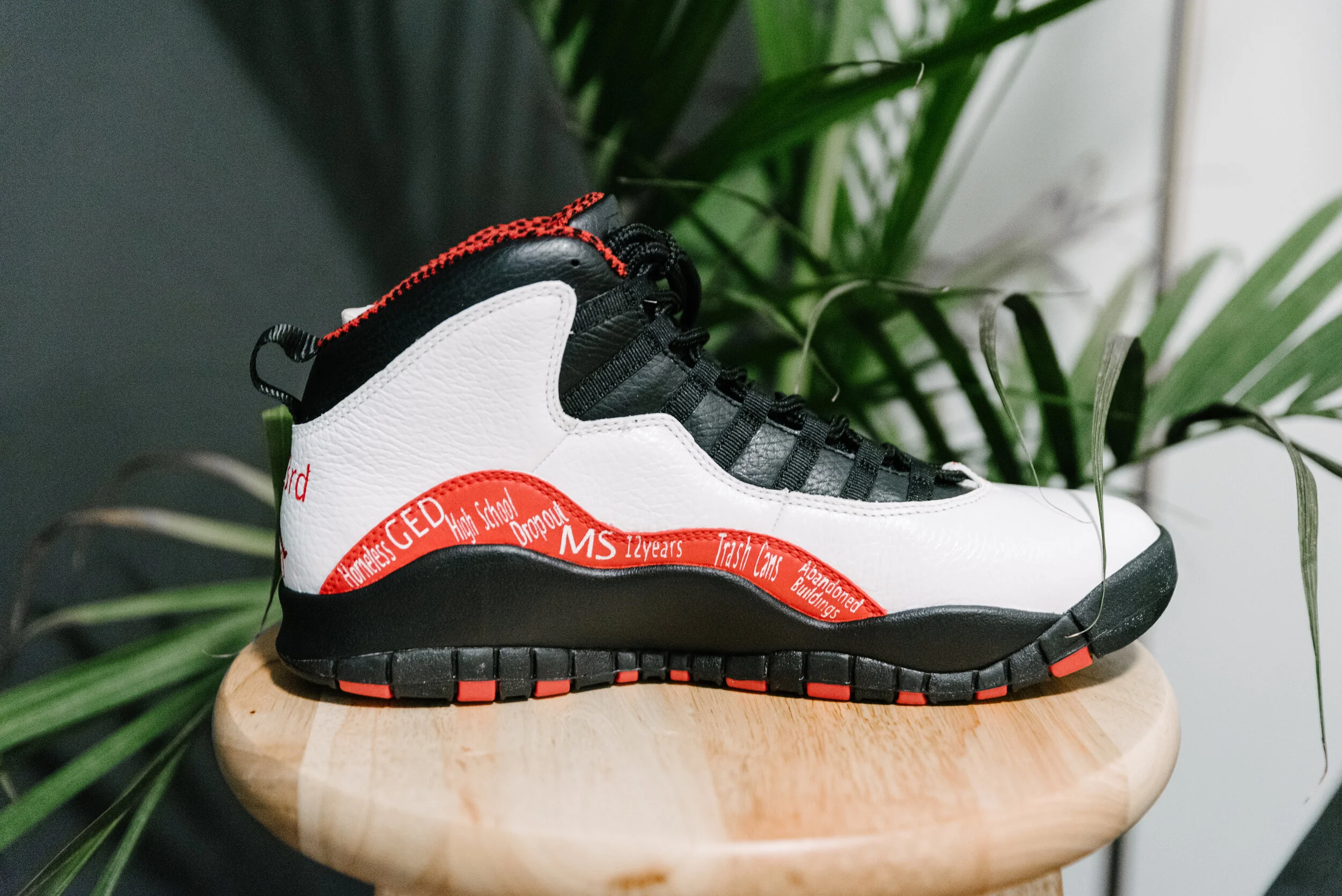 A white, black, and red sneaker placed on a wooden surface, with green plants in the background.