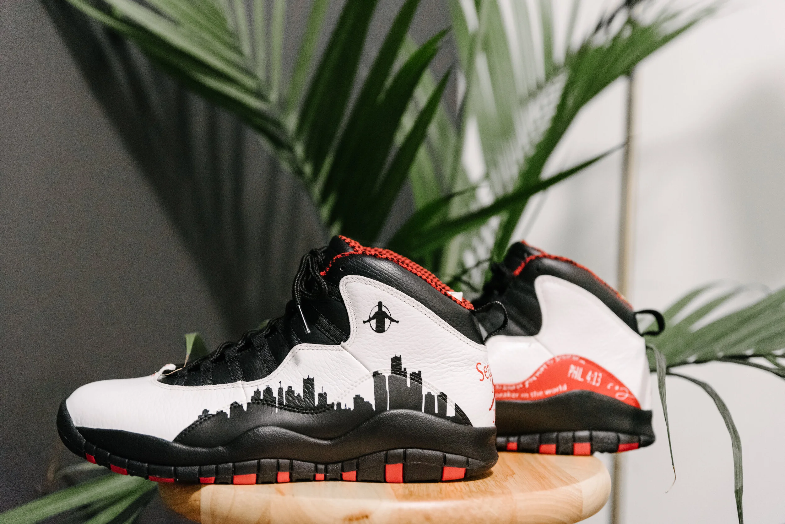 A pair of black and white sneakers with red accents, featuring city skyline and New York City lettering, placed on a wooden surface with green tropical plants in the background.