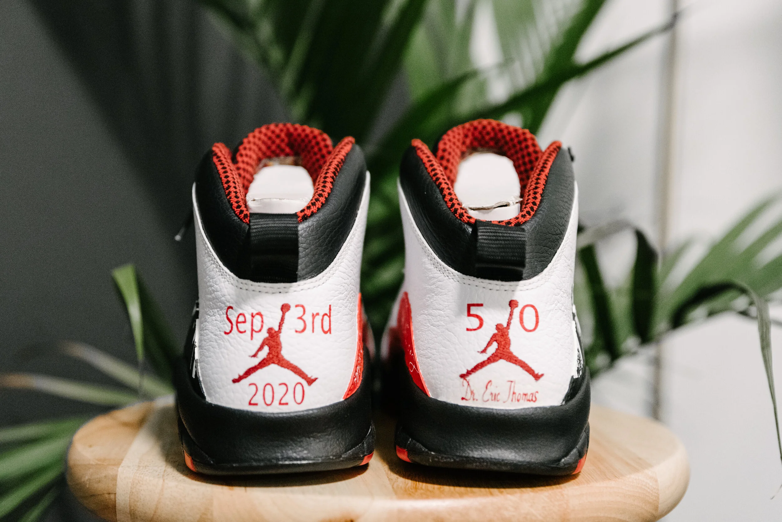 A pair of custom-designed white and black sneakers with red accents, displayed on a wooden surface. The back of the shoes features embroidered details including a silhouette of a basketball player, date, and text, indicating a memorial or tribute. Th