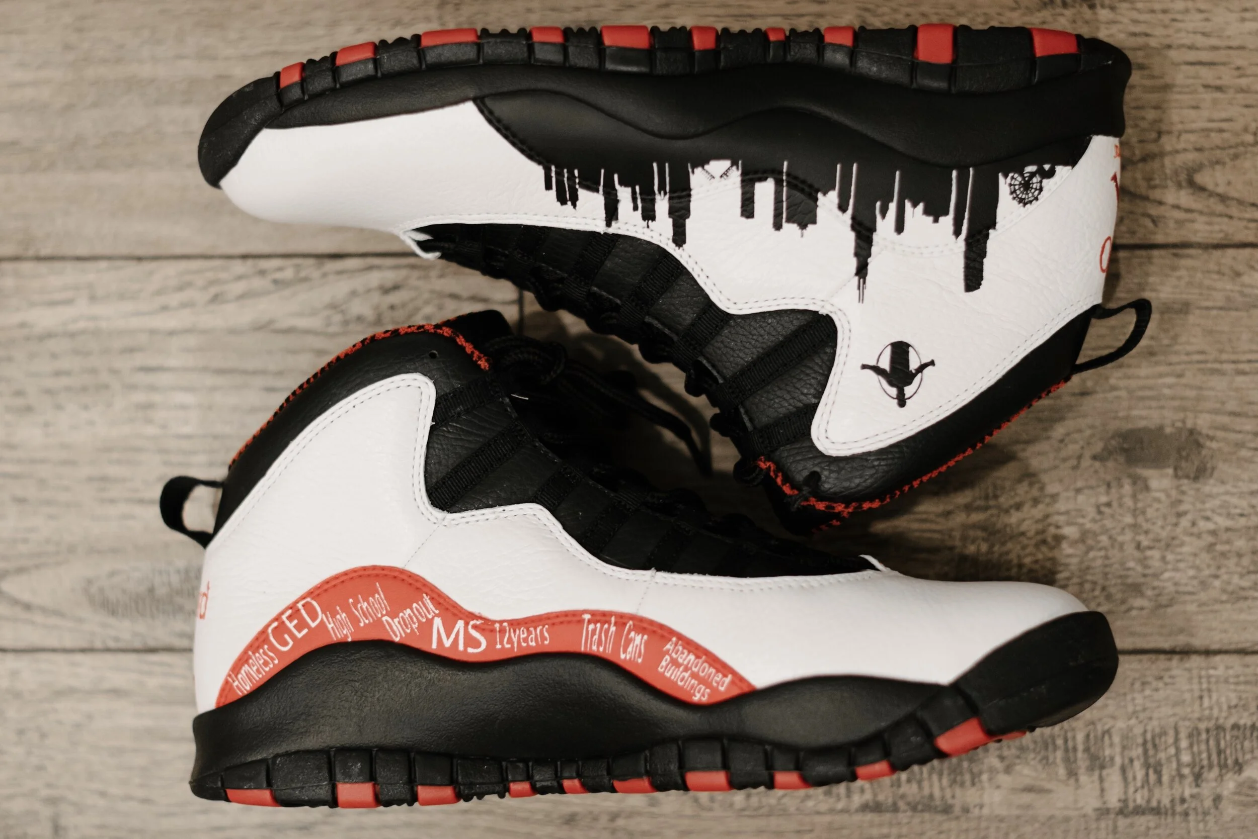 Pair of custom-designed black, white, and red sneakers with cityscape graphics and text elements. The shoes feature a white upper with black accents and red details on the soles and lace areas, displayed on a wooden surface.