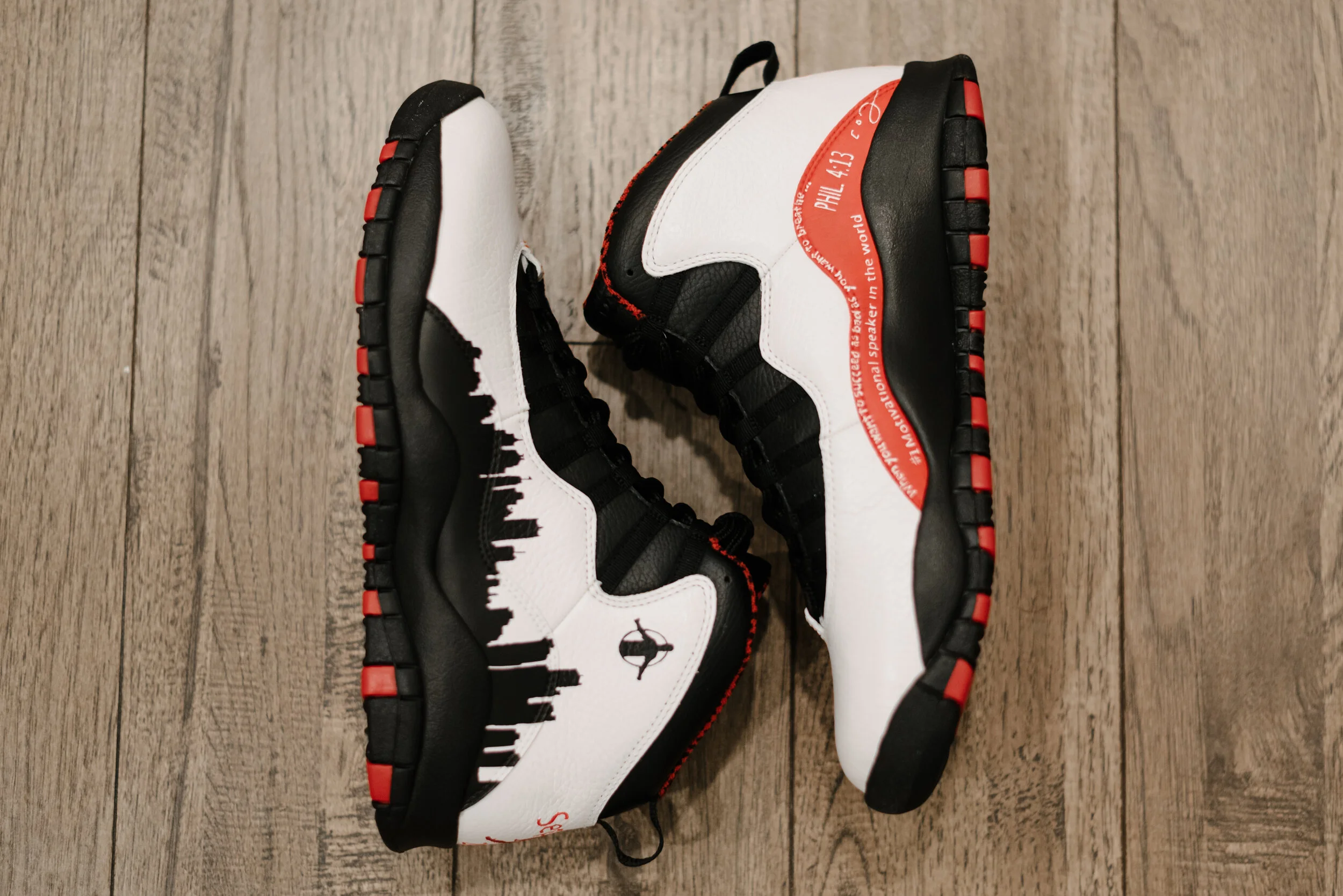 Pair of limited-edition sneaker shoes with black, white, and red design, featuring skyline silhouette and text, on wooden floor.