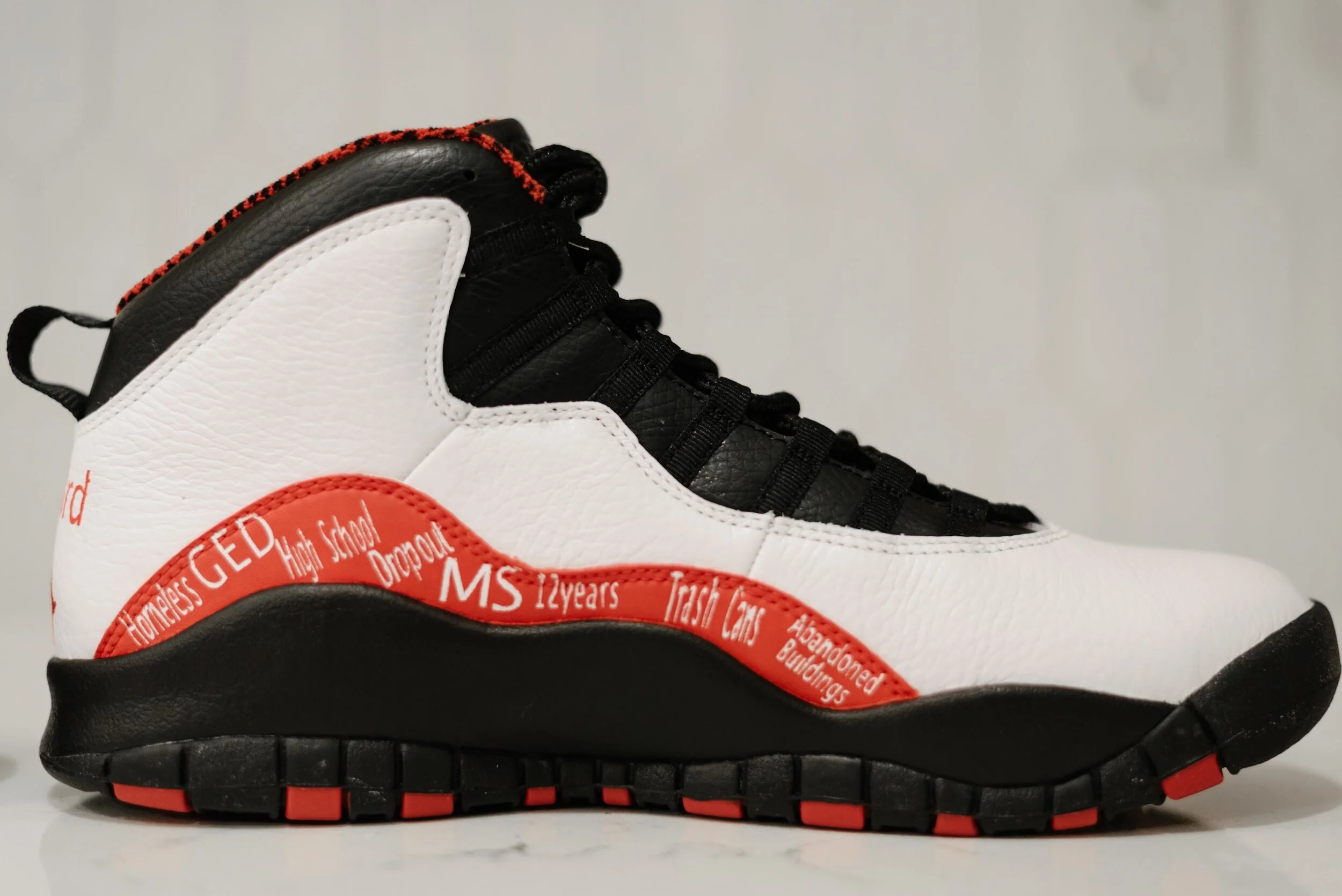 A white, black, and red sneaker with black laces and a black sole, featuring handwritten-style words and phrases in red around the midsole.