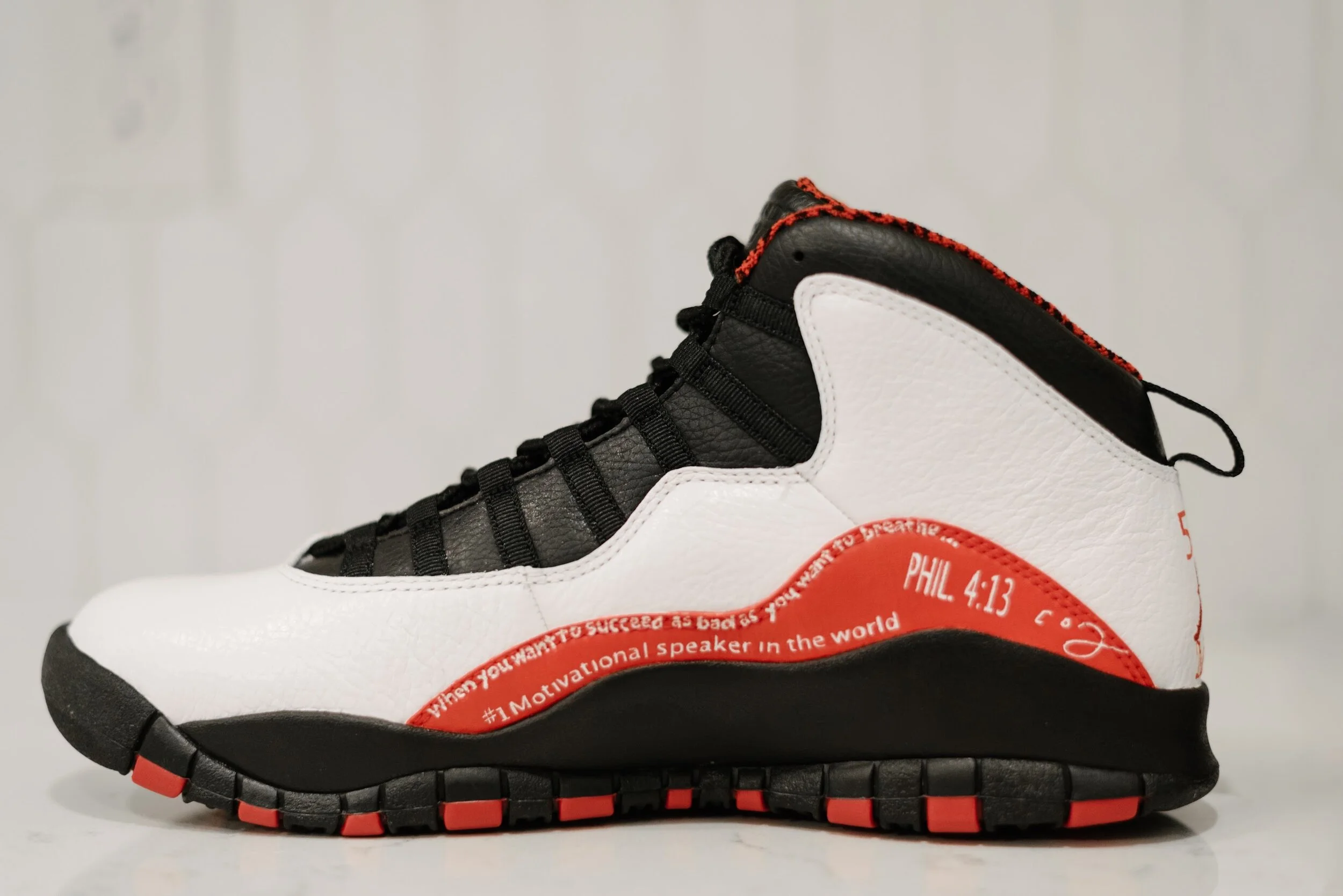 A close-up of a black, white, and red athletic shoe with motivational quotes and scripture, and a distinctive sole design.