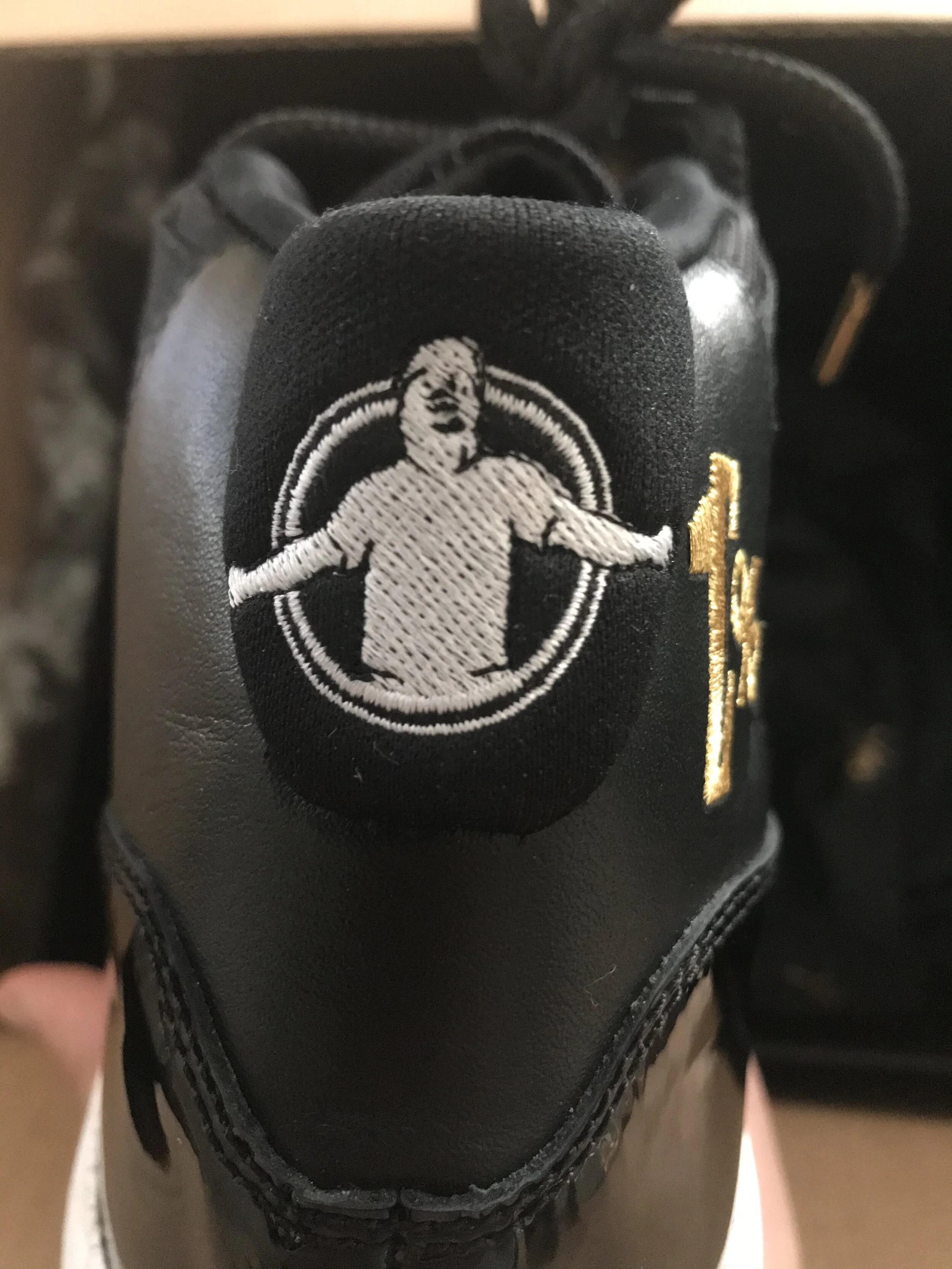 Close-up view of the tongue of a black sneaker, embroidered with a white monkey with arms outstretched inside a circle and a gold number '13' on the side.