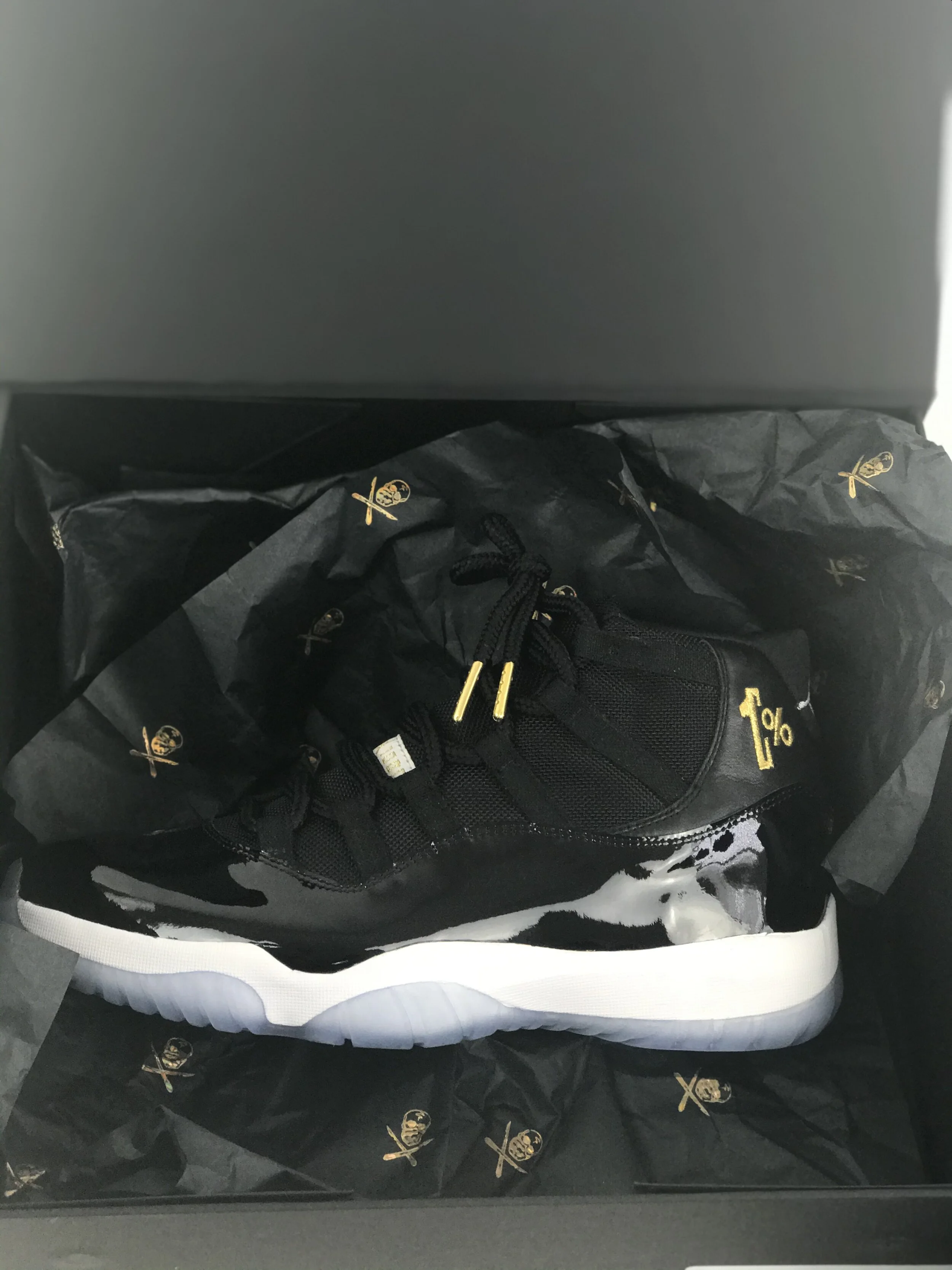 Black sneaker with white sole inside a box, lined with black paper with gold skull and crossbones logo.