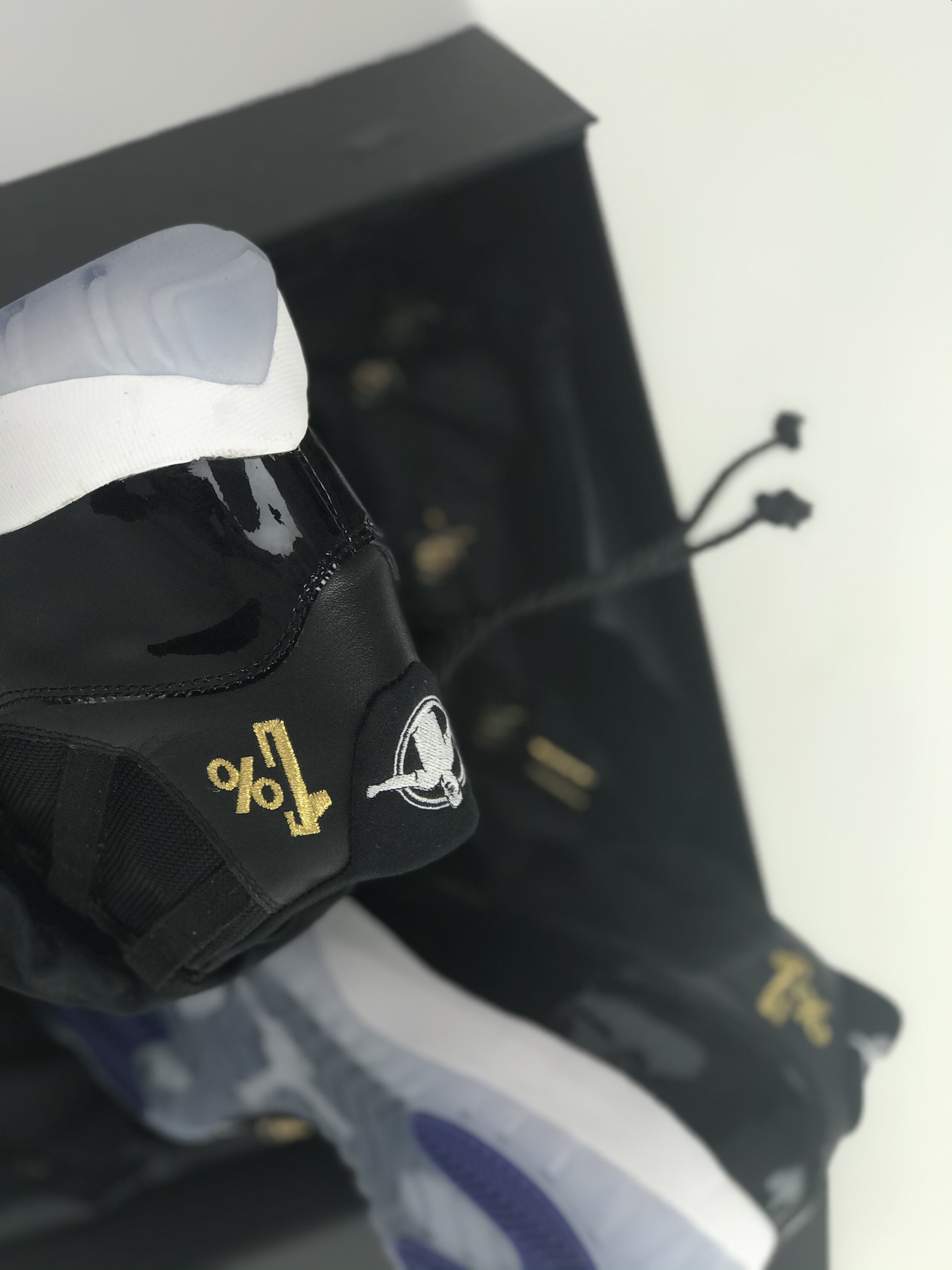 Close-up of a black racing glove with yellow percentage symbol and logo, placed on a white and purple racing glove, with a black box or case in the background.