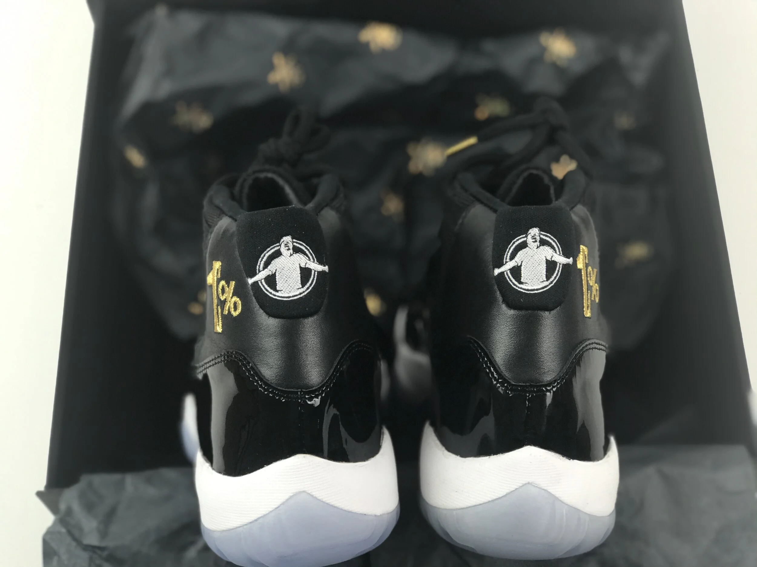 Pair of black and white sneakers with embroidered gold "1%" and a white figure on the tongue, placed on satin fabric inside a box.