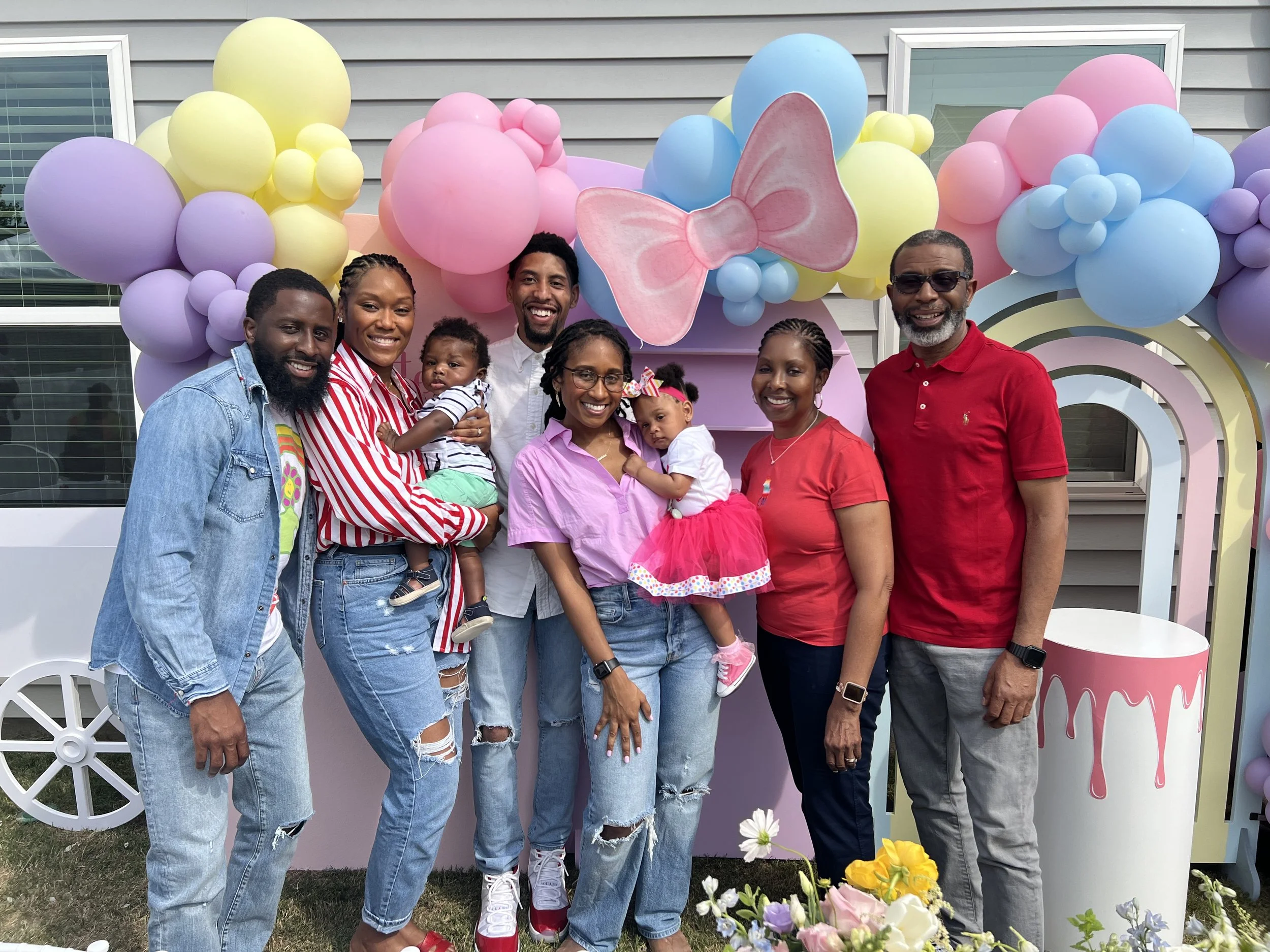 Family and friends celebrating at a colorful party with pastel balloon arch, pink bow decoration, and flower arrangements.