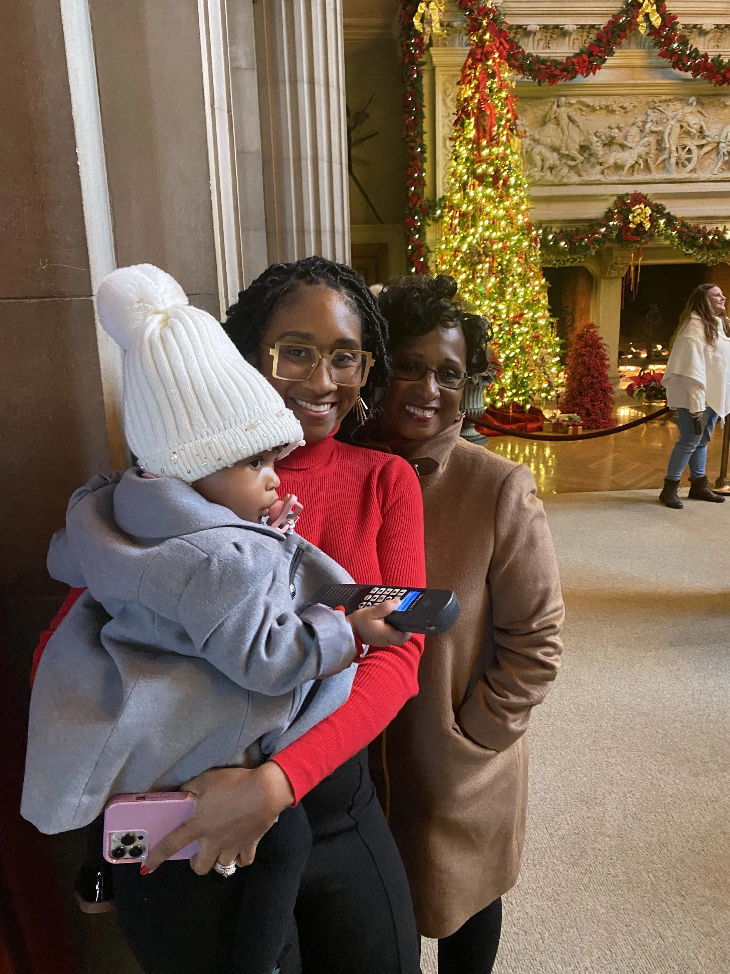 Three people, two women and a young child, smiling indoors near a decorated Christmas tree. The woman on the left is holding a phone, and the child is wearing a white knit hat and holding a remote control.