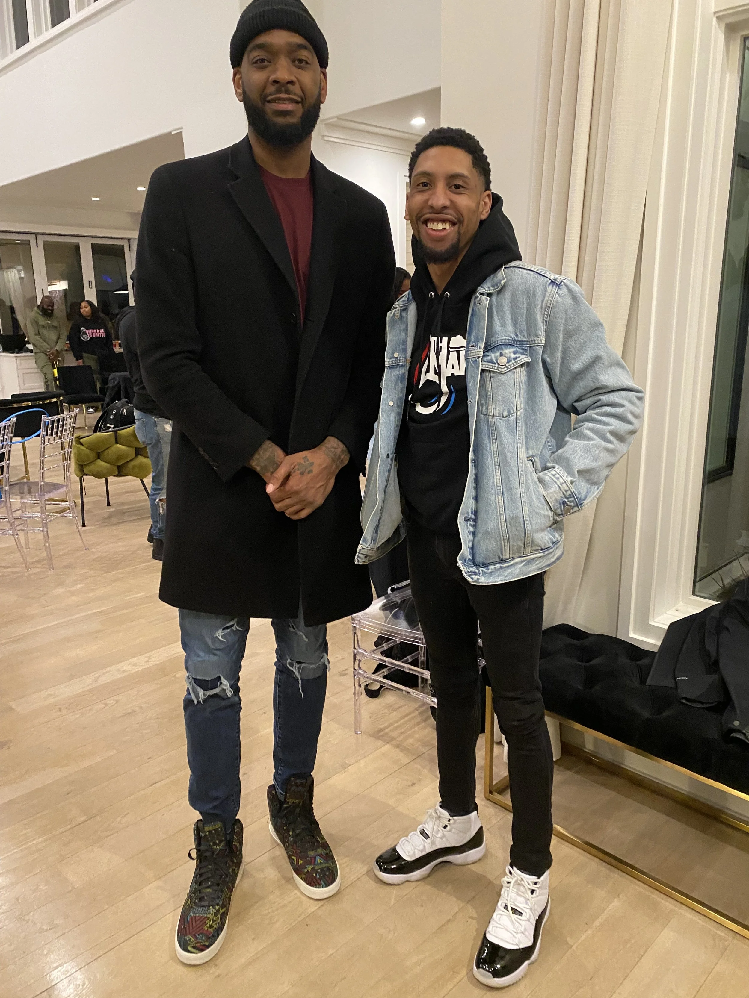 Two men standing indoors, smiling at the camera. One is tall, wearing a black coat, jeans, colorful sneakers, and a black beanie. The other is shorter, wearing a denim jacket over a hoodie, black pants, and white and black sneakers.