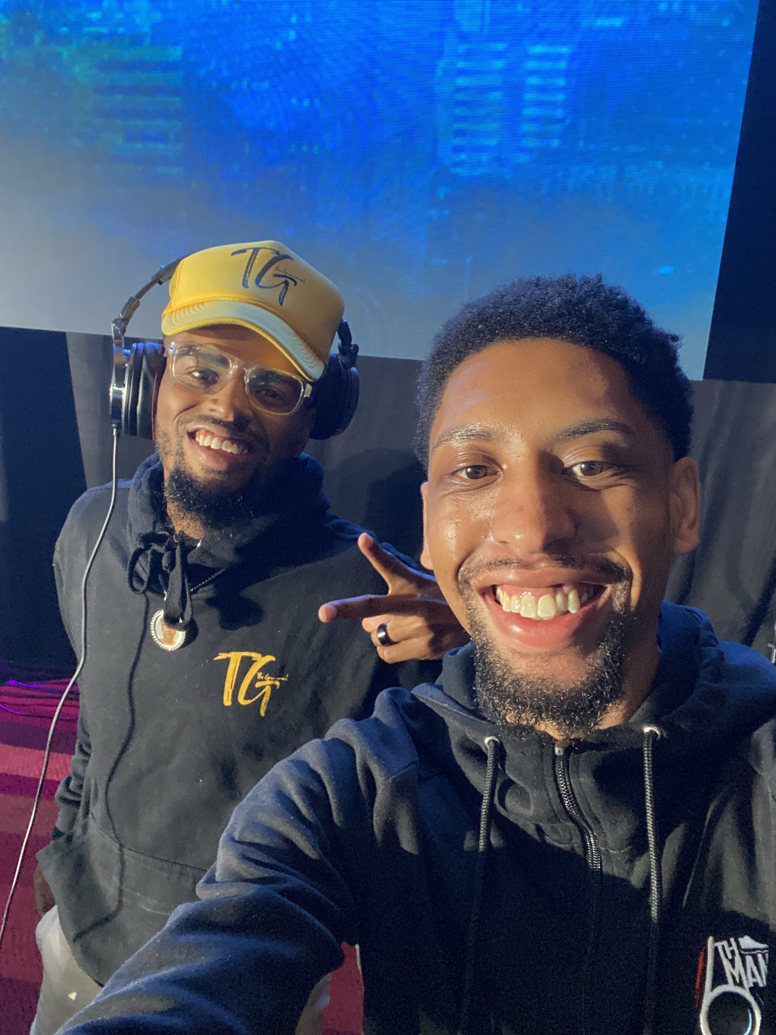 Two men smiling and posing for a selfie in front of a large screen, one wearing headphones and glasses, the other showing a peace sign, both dressed in black hoodies.