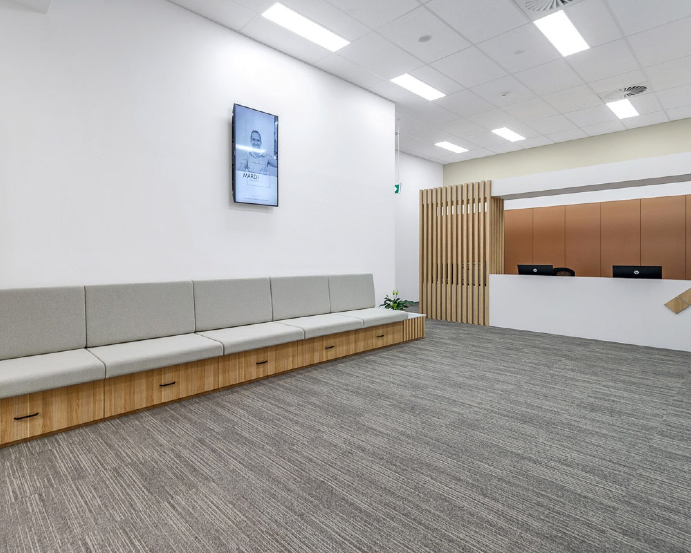Modern waiting area with gray cushioned seating, a digital screen on the wall, a wooden reception desk, and gray carpet flooring.