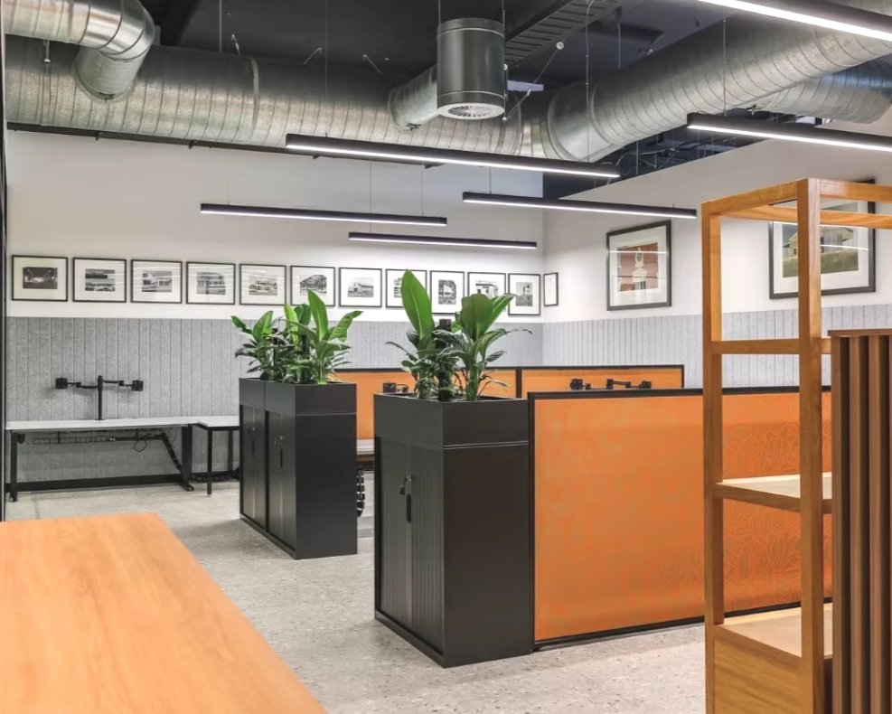 Modern open office space with orange dividers, black cabinets, and green plants. Industrial ceiling with exposed ductwork and framed artwork on walls.