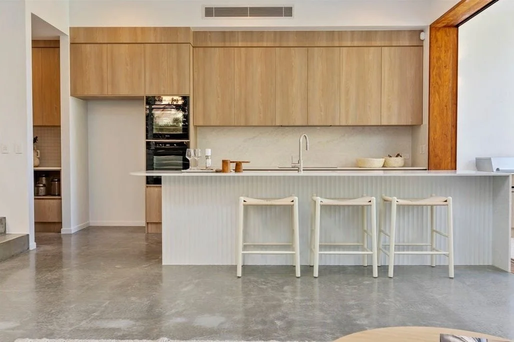Modern kitchen with wooden cabinets, a white island with three bar stools, and stainless steel appliances on a grey floor.