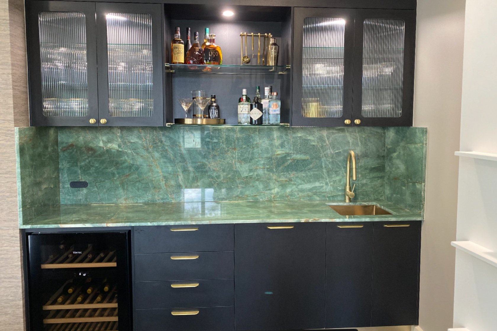 Home bar area with dark cabinets, frosted glass doors, glass shelves holding liquor bottles and glasses, gold sink faucet, green marble countertop and backsplash, and wine fridge.