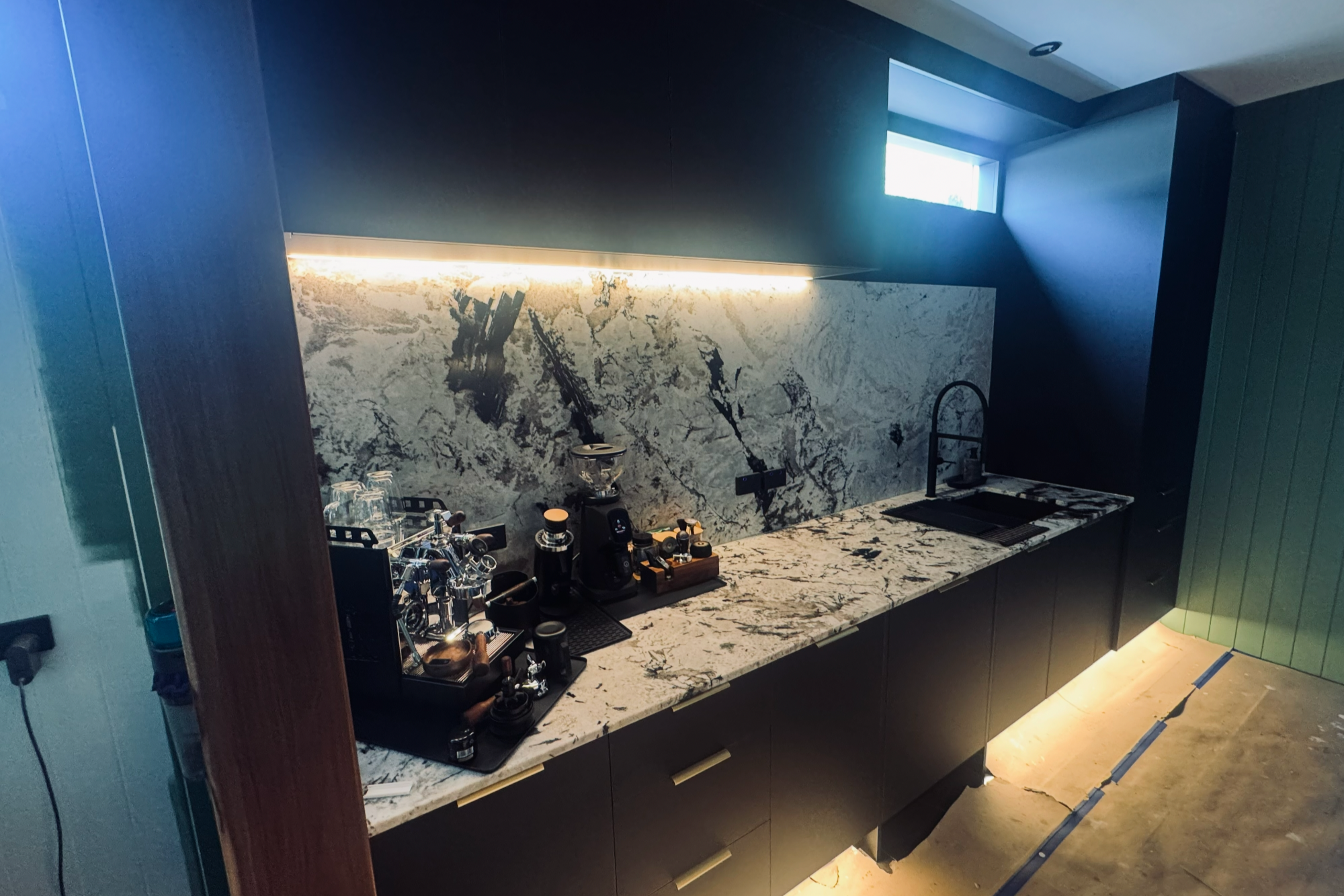 Modern kitchen with black cabinetry, white marble countertop with dark veining, and marble backsplash. Features a sink, coffee station, and under-cabinet lighting. Small window provides natural light.
