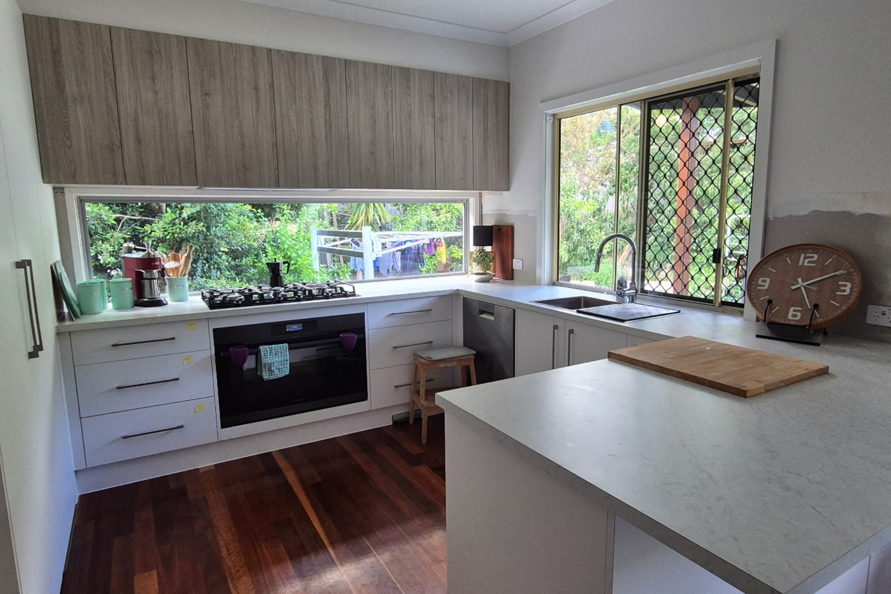 Modern kitchen with wooden cabinets, white countertops, and hardwood floors. Outside greenery visible through large windows. Contains kitchen appliances, a clock, and a cutting board on the counter.