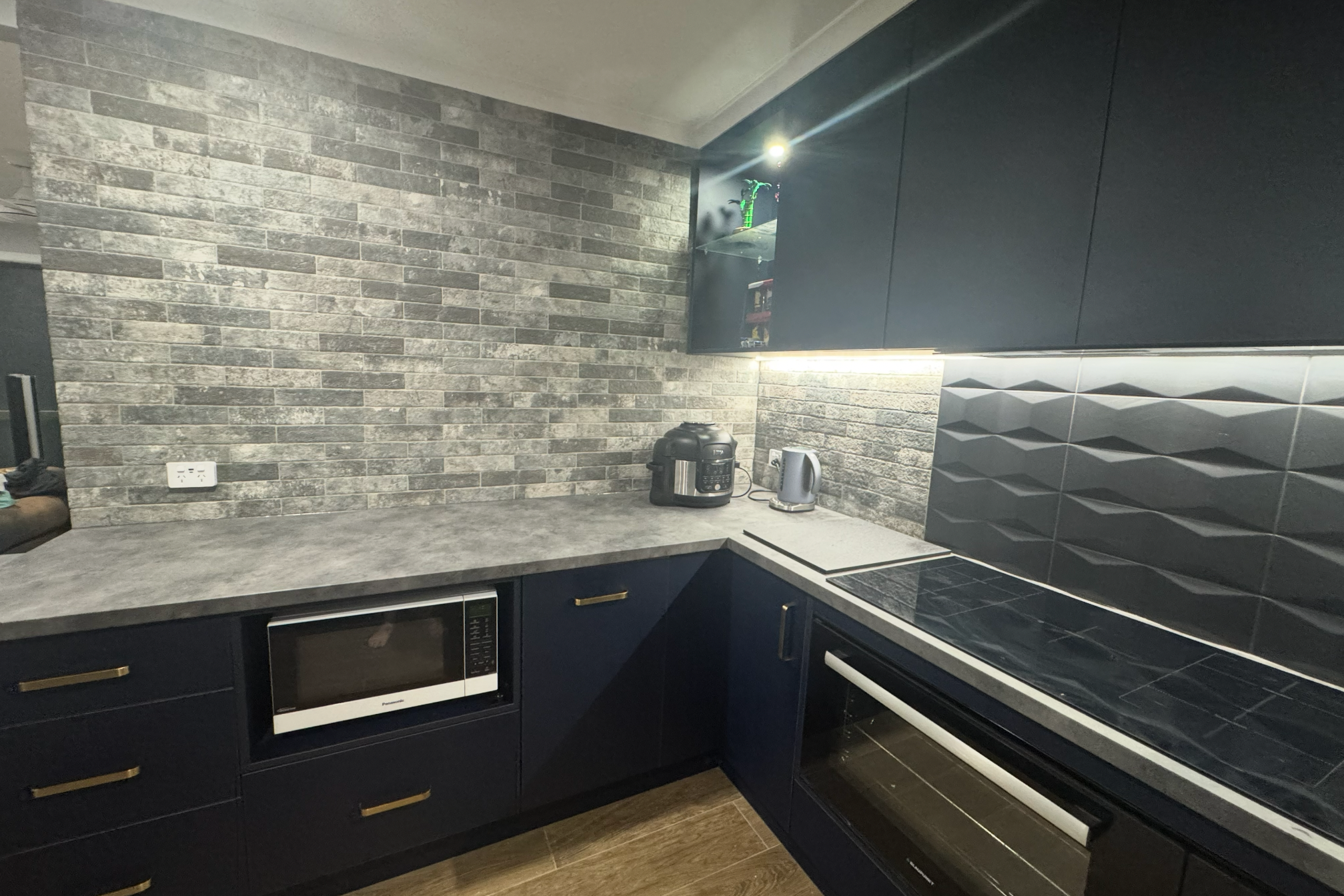 Modern kitchen with grey brick backsplash, dark blue cabinets with gold handles, and black appliances including a microwave and oven. Countertop holds a rice cooker, kettle, and a small potted plant.