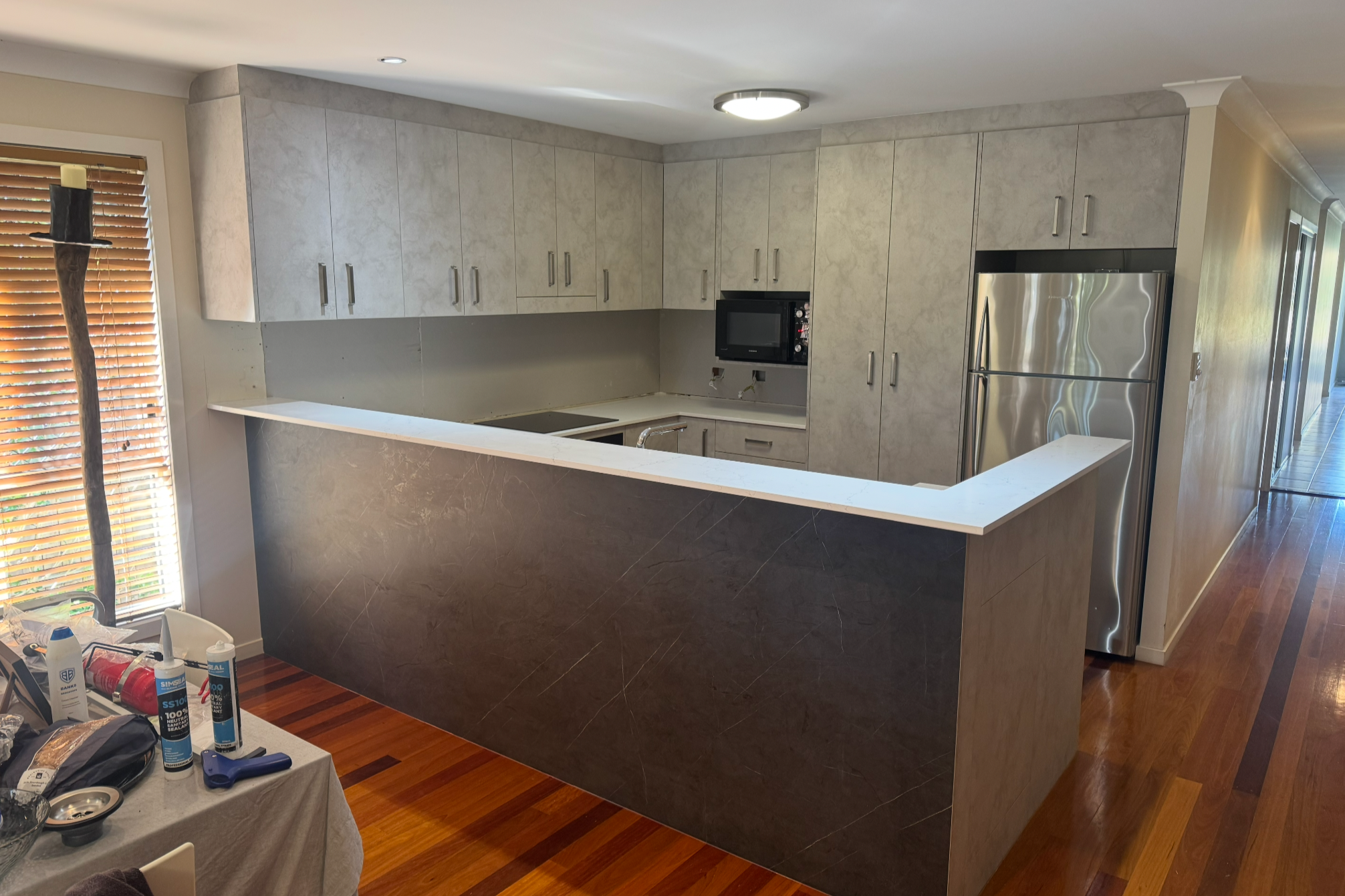 Kitchen with gray cabinets, stainless steel refrigerator, microwave, and black countertops, overlooking a hallway with hardwood floors.