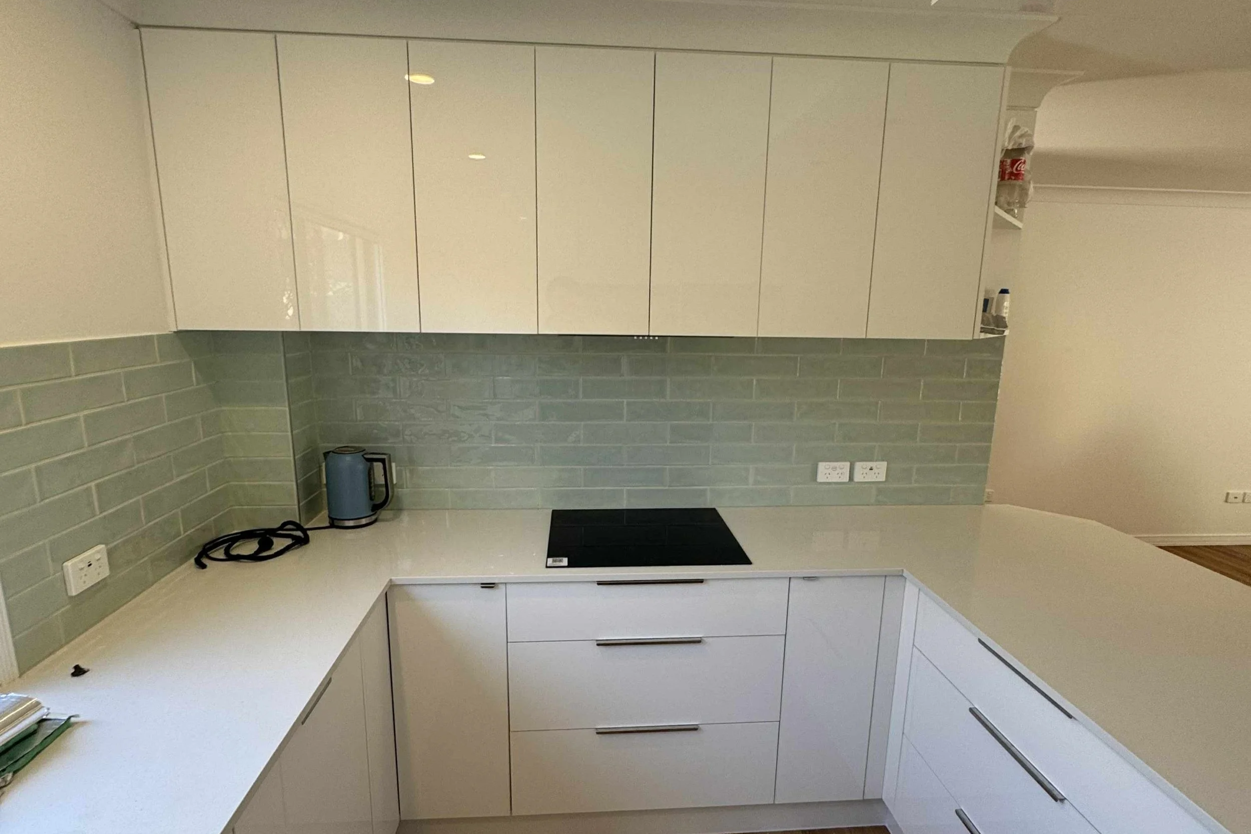 Modern kitchen with white cabinets, light green tile backsplash, electric stovetop, kettle, and outlets.
