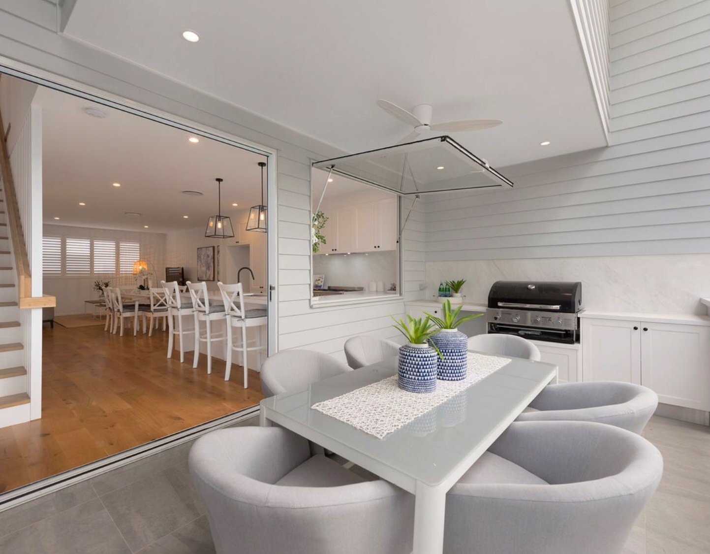 Modern indoor-outdoor dining area with a glass table, gray chairs, decorative vases, built-in barbecue, and wooden flooring. Interior features a long dining table with white chairs, ceiling lights, and staircase.