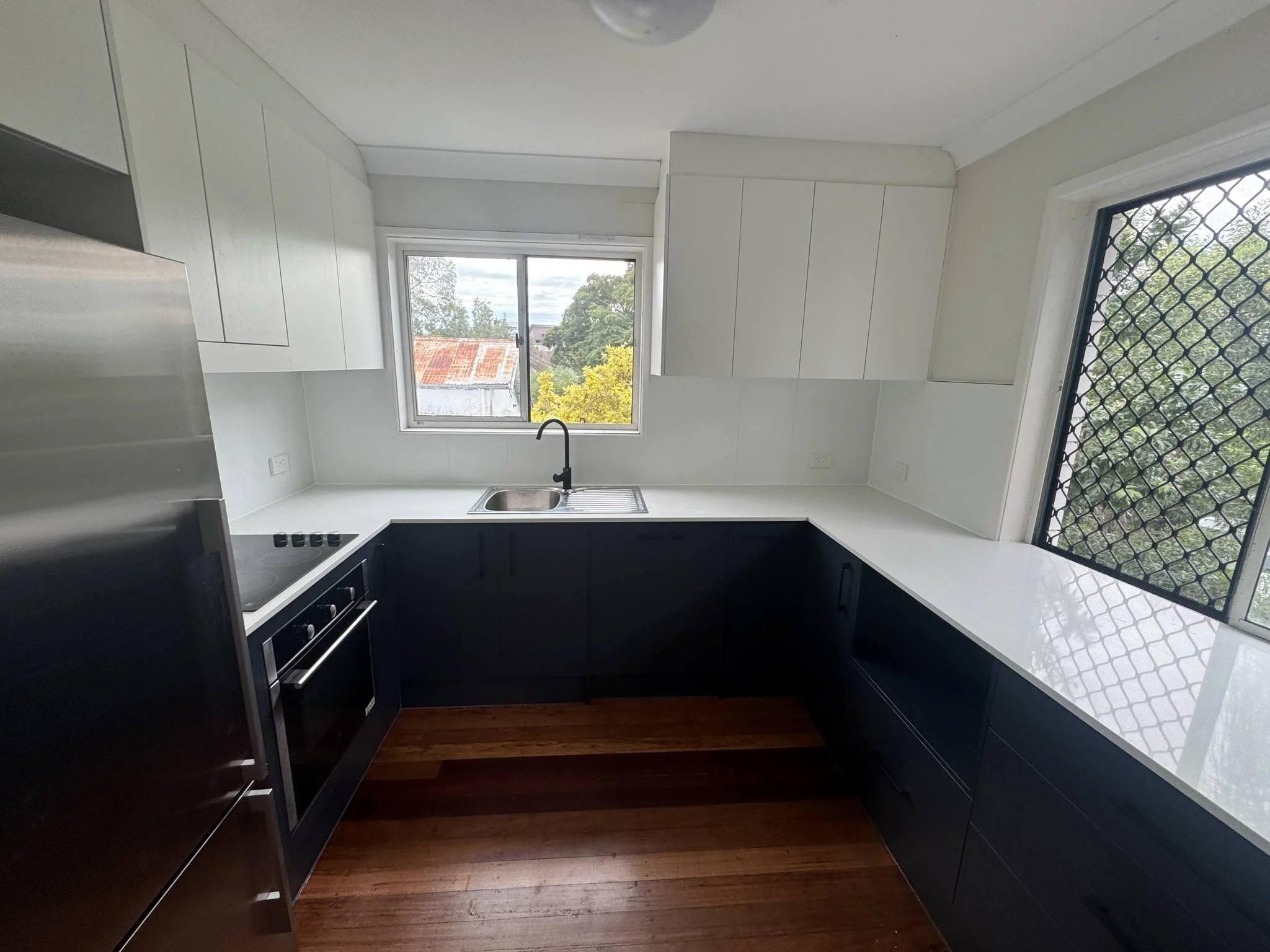 Modern kitchen with black lower cabinets, white upper cabinets, a stainless steel oven, black cooktop, and a window above the sink with a view of trees and rooftops.