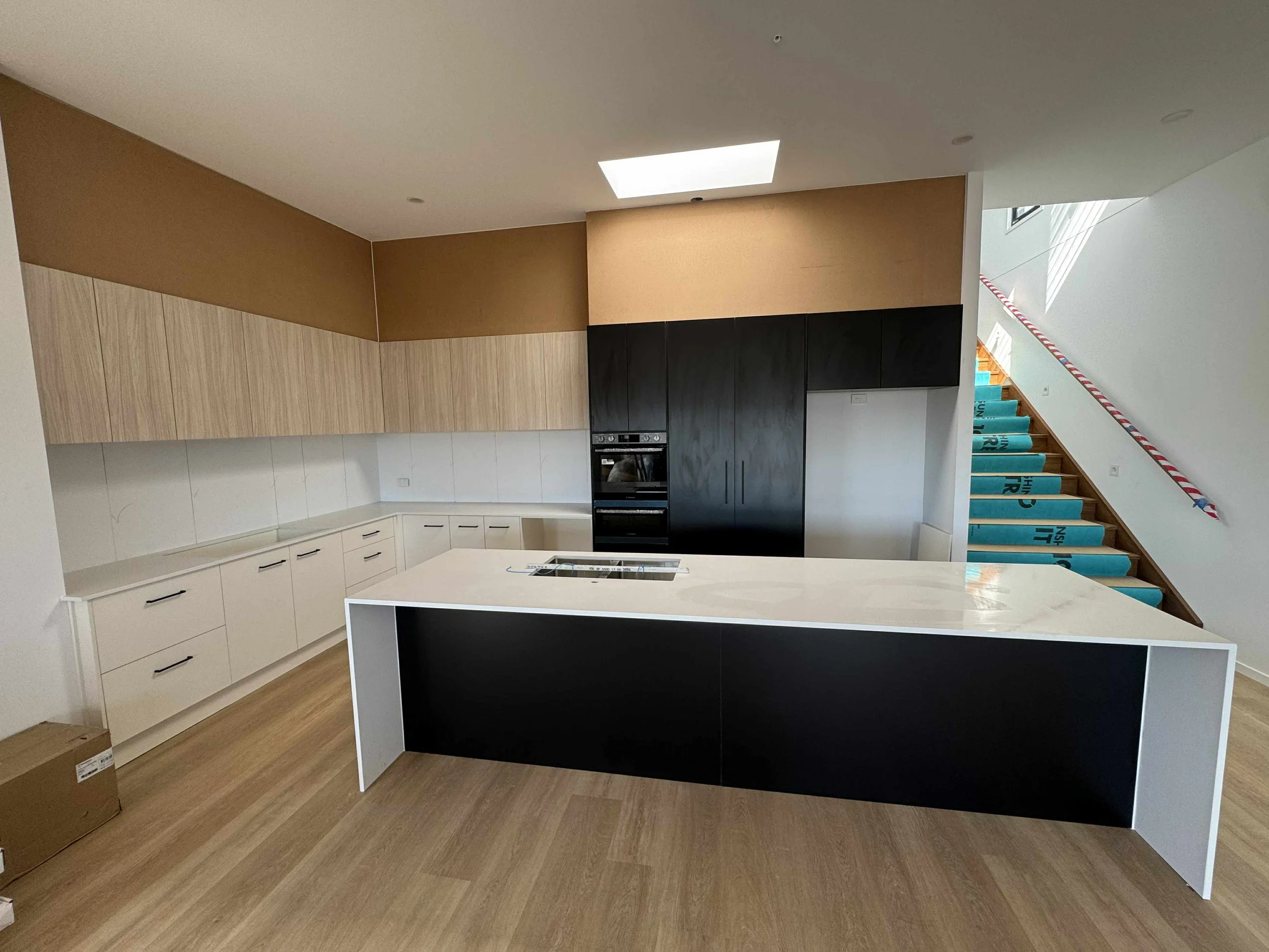 Modern kitchen with white countertops, black and wood cabinets, an island with a built-in stove, and an staircase with blue non-slip treads and red-and-white striped handrail.