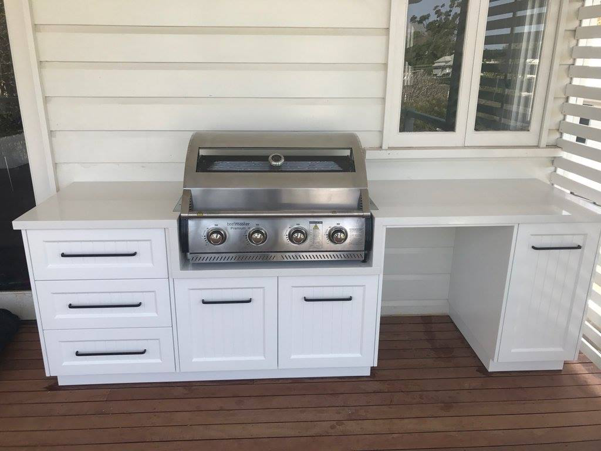 Outdoor kitchen setup with a built-in stainless steel grill, white cabinets, and drawers on a wooden deck.