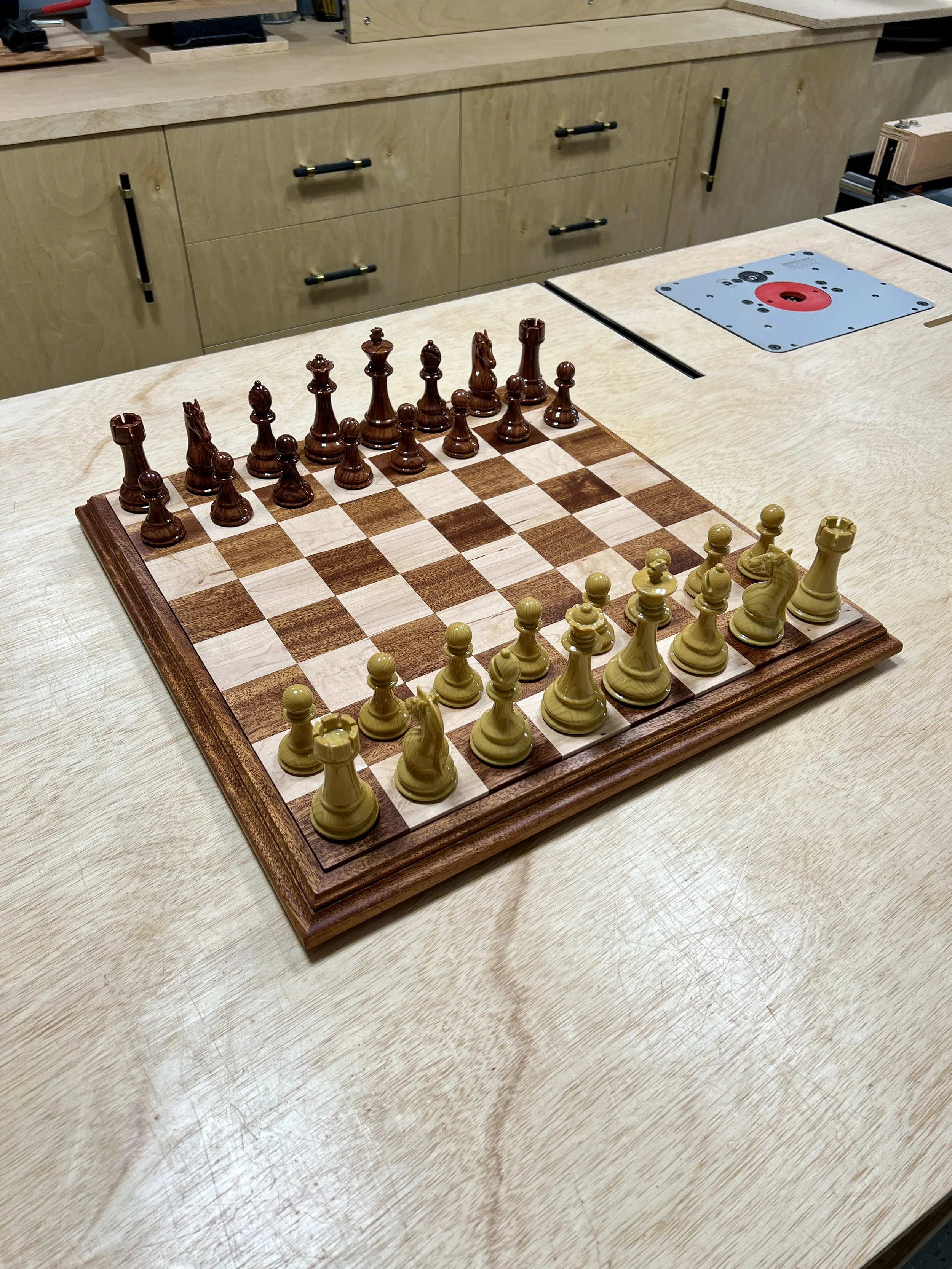 Wooden chessboard with chess pieces set up for a game, placed on a light-colored table in a workshop or woodworking space.