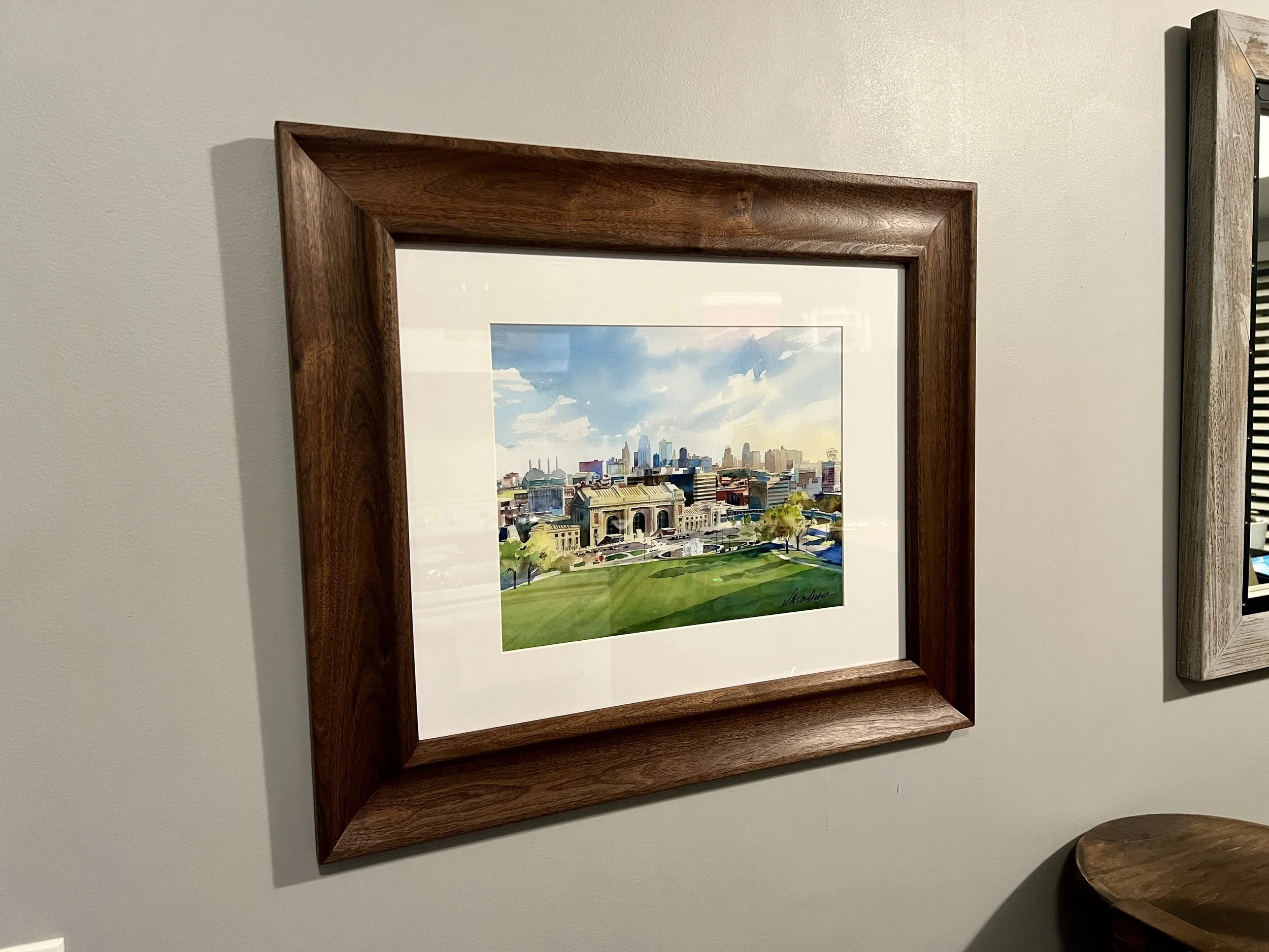 A colorful watercolor painting of a cityscape with a large building in the foreground, green lawns, and tall skyscrapers in the background, framed in a wooden frame, hanging on a beige wall.