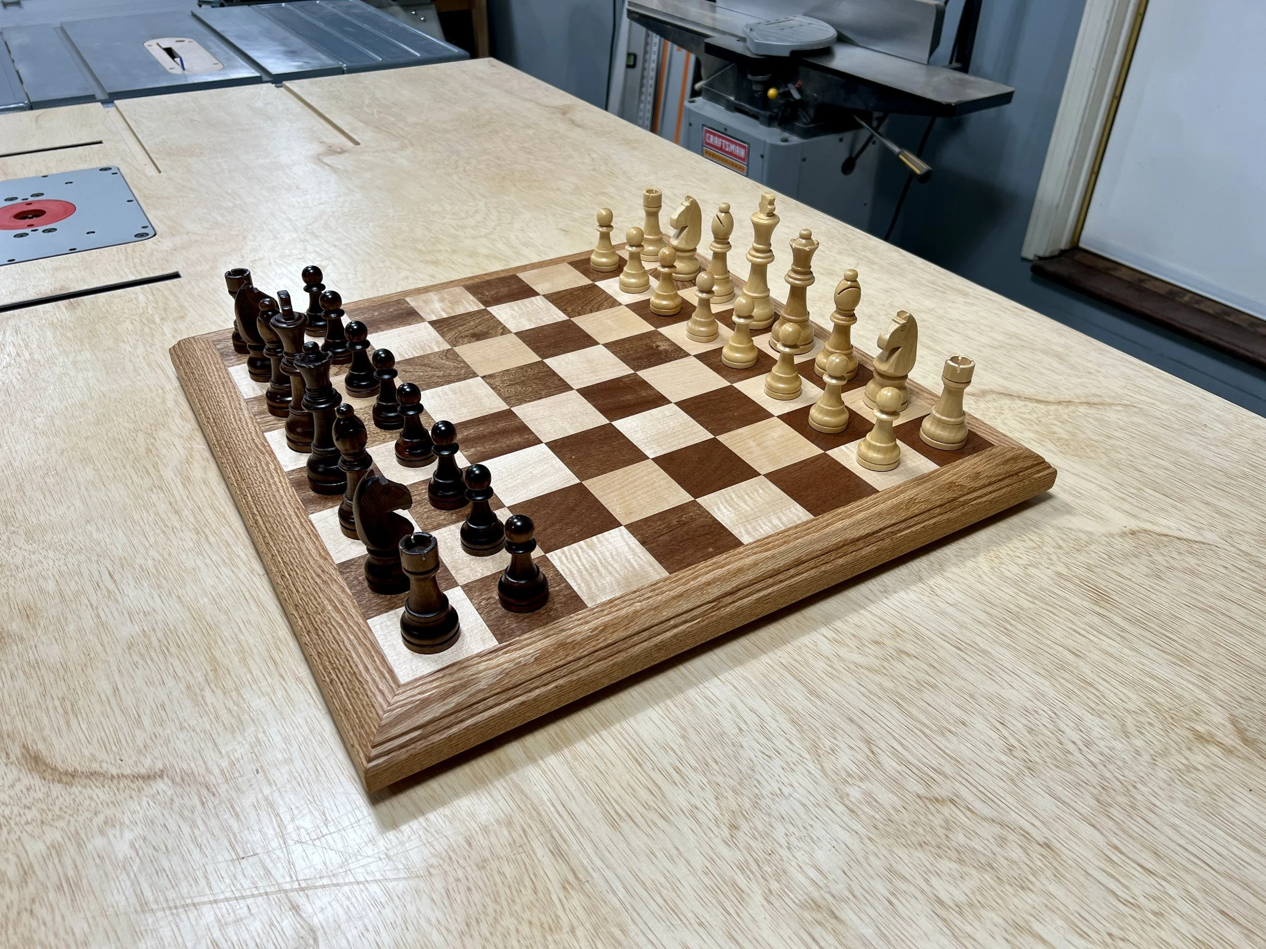 A chessboard with black and white pieces set up in starting positions, on a wooden table in a workshop.