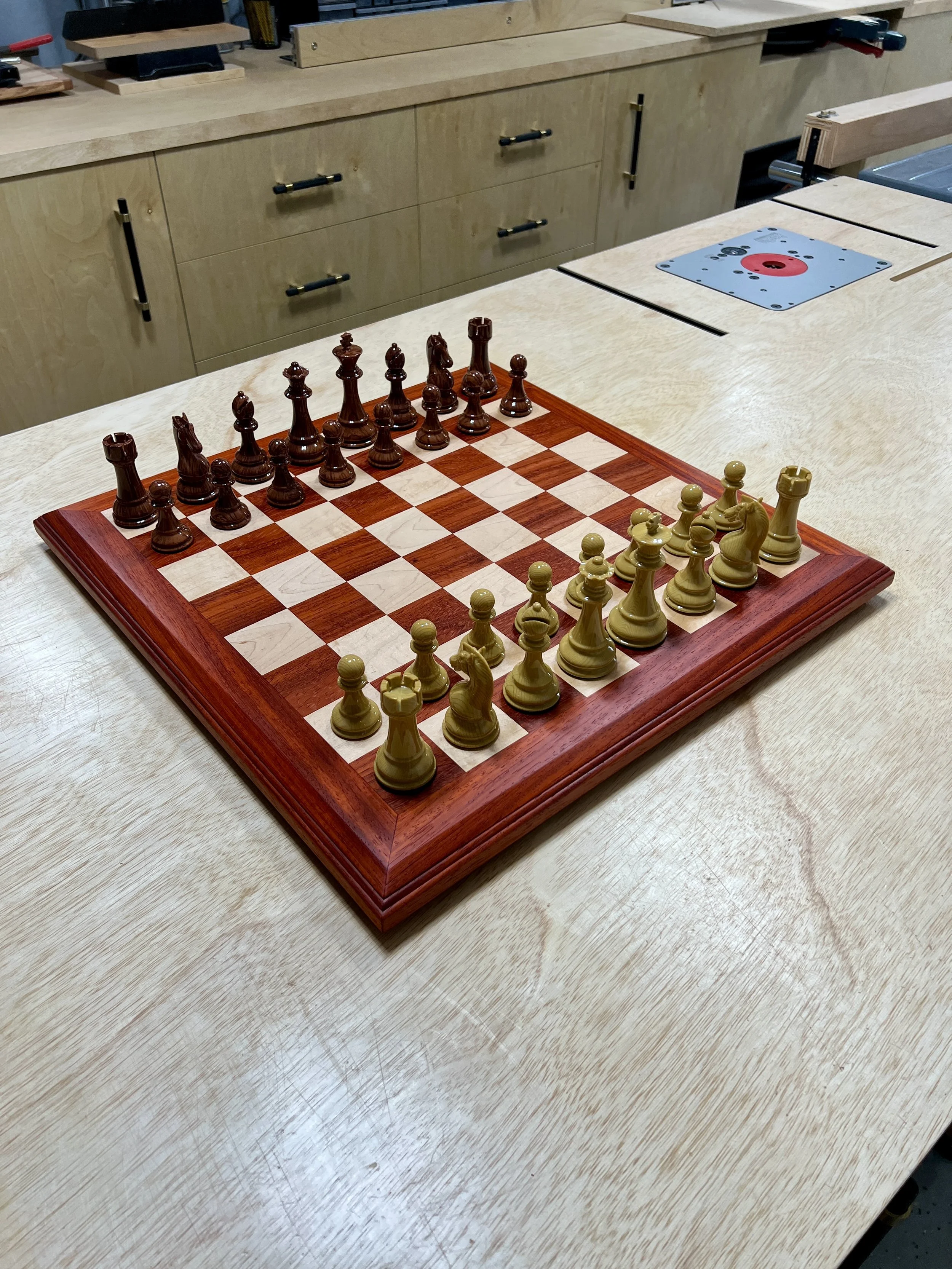 Chessboard with wooden pieces set up for a game on a light-colored work table in a workshop.