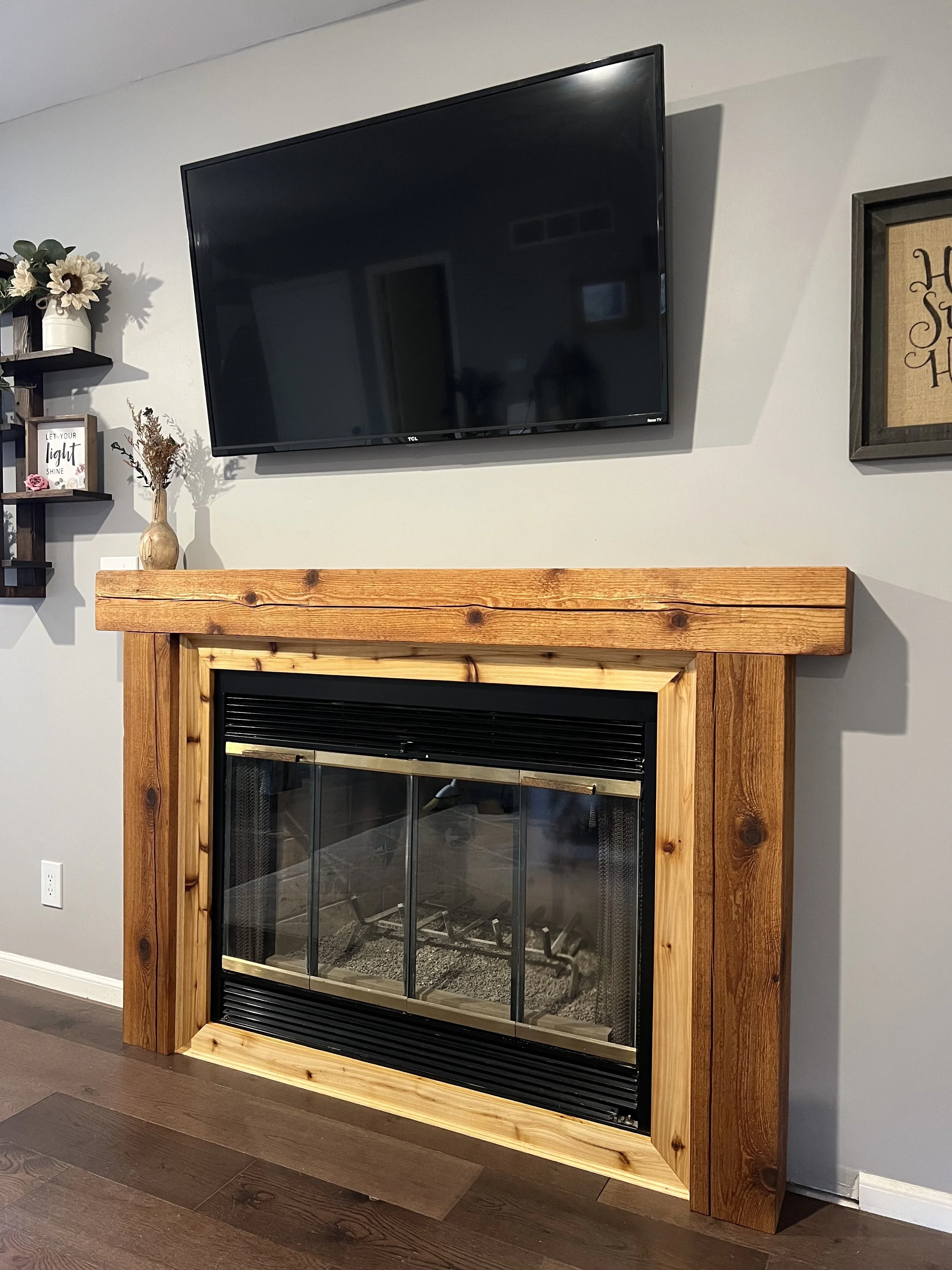 Living room fireplace with a wood mantel, with a flat-screen TV mounted above, and decorative items including a vase with dried flowers and framed artwork on the wall.