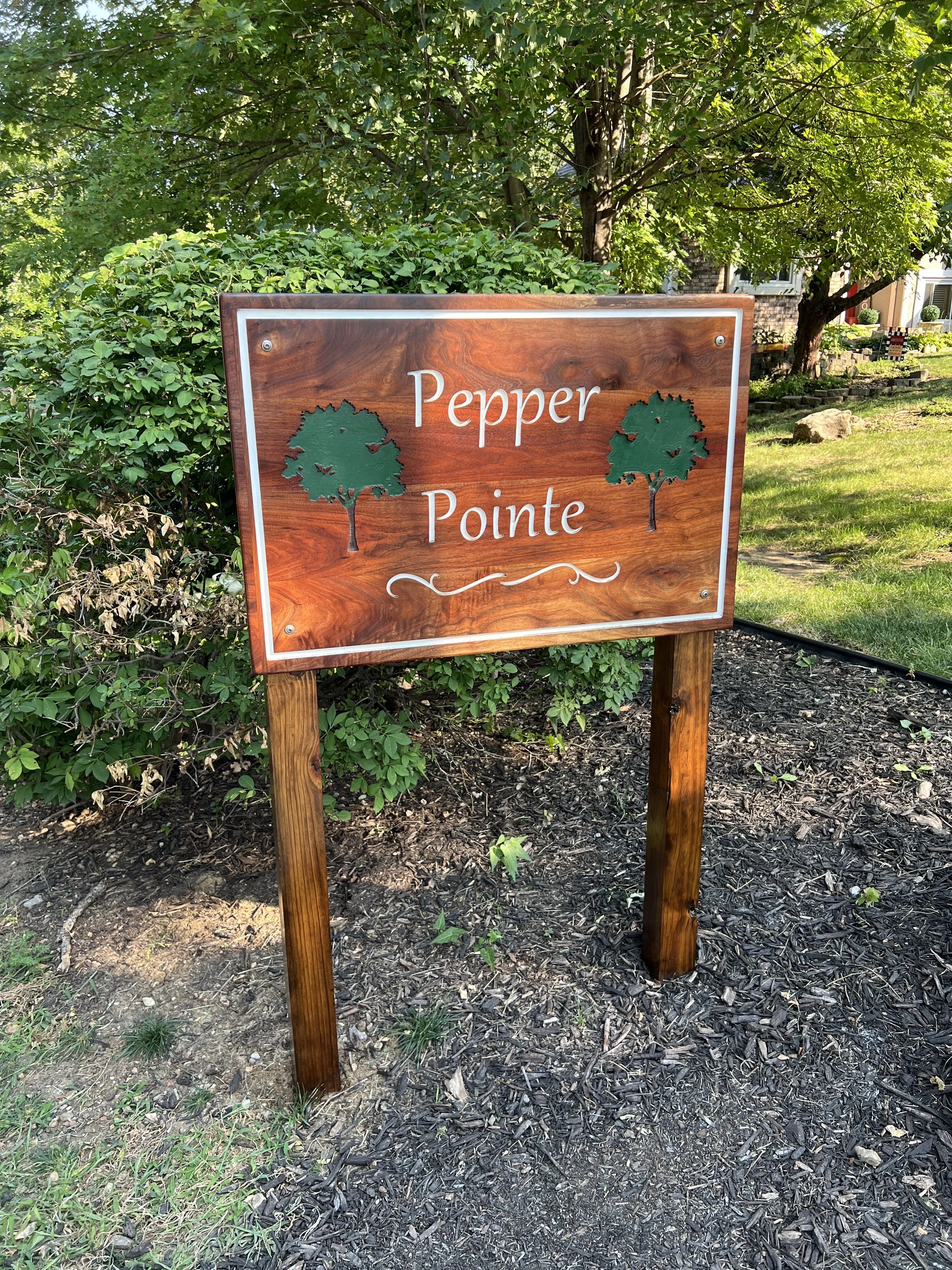 Wooden sign with green trees and white text that reads "Pepper Pointe," situated in a garden with bushes and trees in the background.