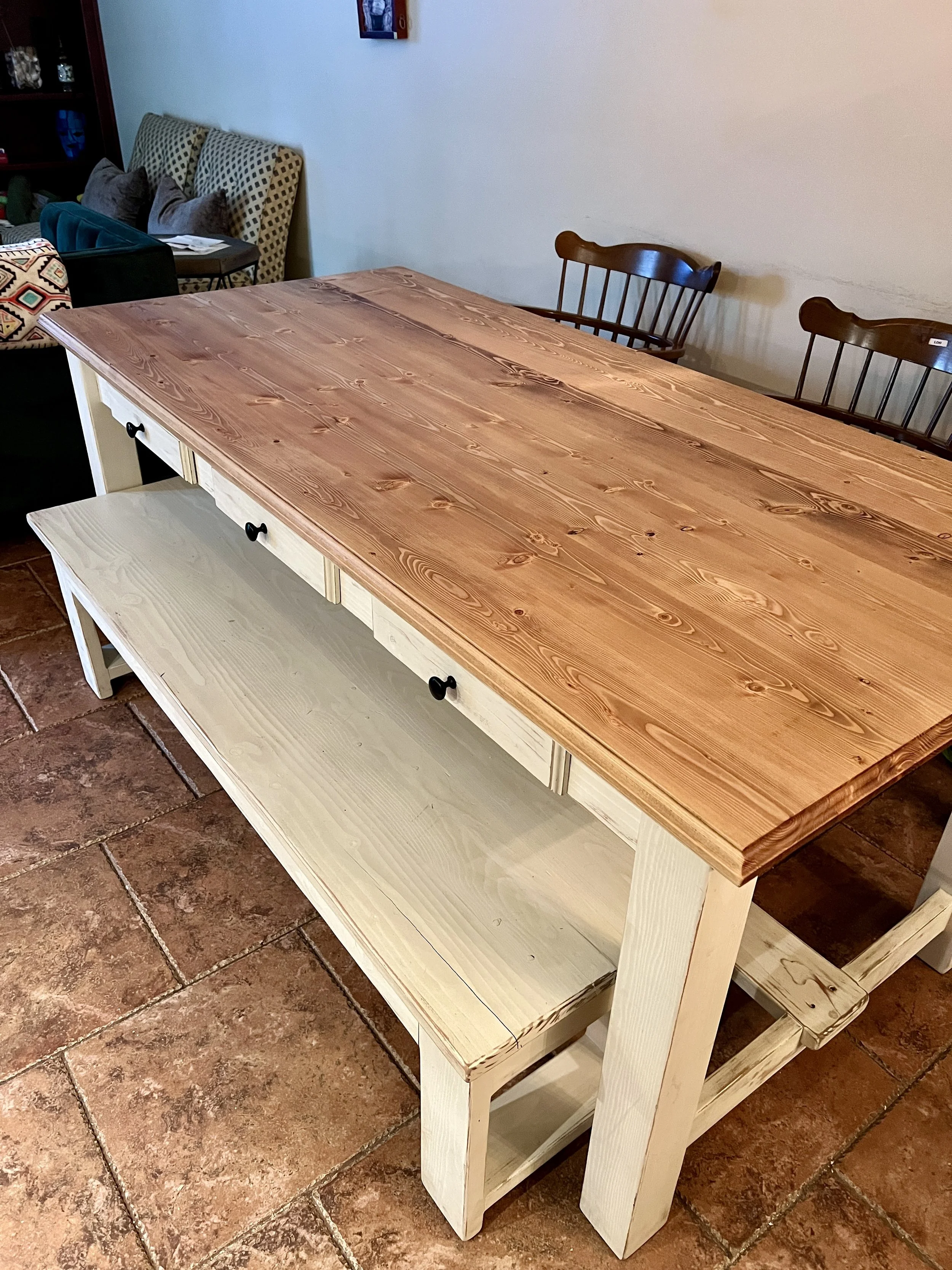 A wooden dining table with a natural wood top, a white base with black knobs, and a lower shelf, set on a tiled floor in a room with furniture and cushions in the background.
