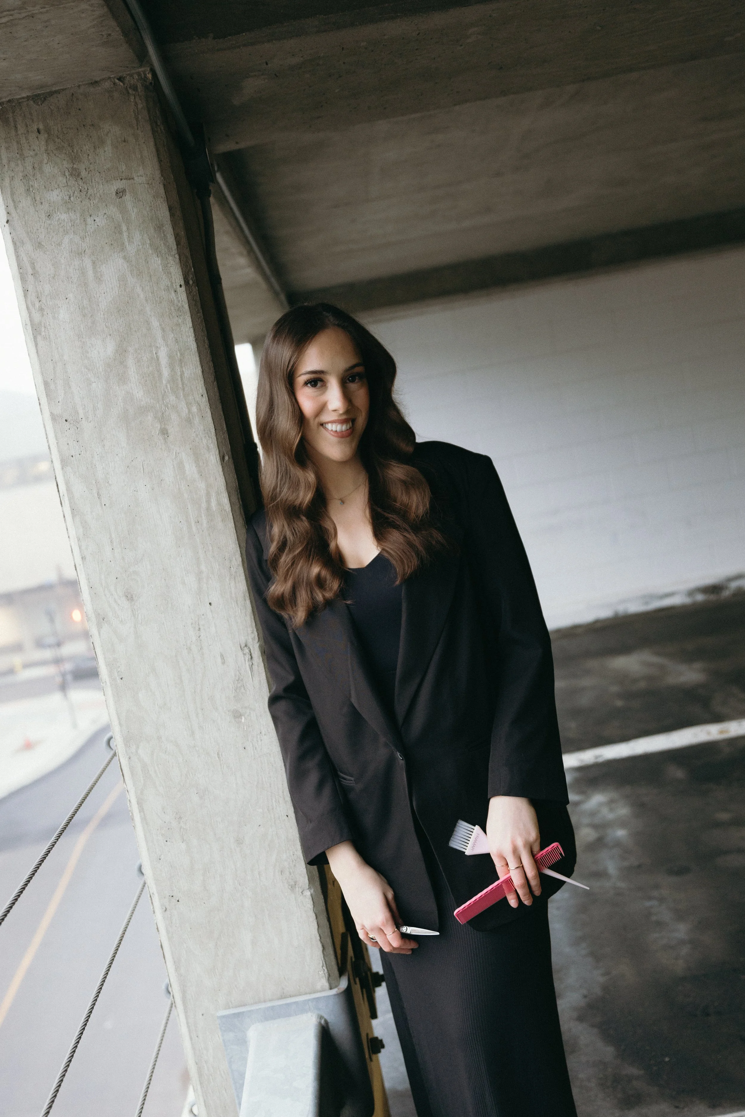 A young woman with long wavy brown hair, wearing a black blazer and black dress, stands leaning against a concrete pillar in an indoor parking garage. She is smiling, holding a pink hairbrush, a comb, and a small pair of scissors.