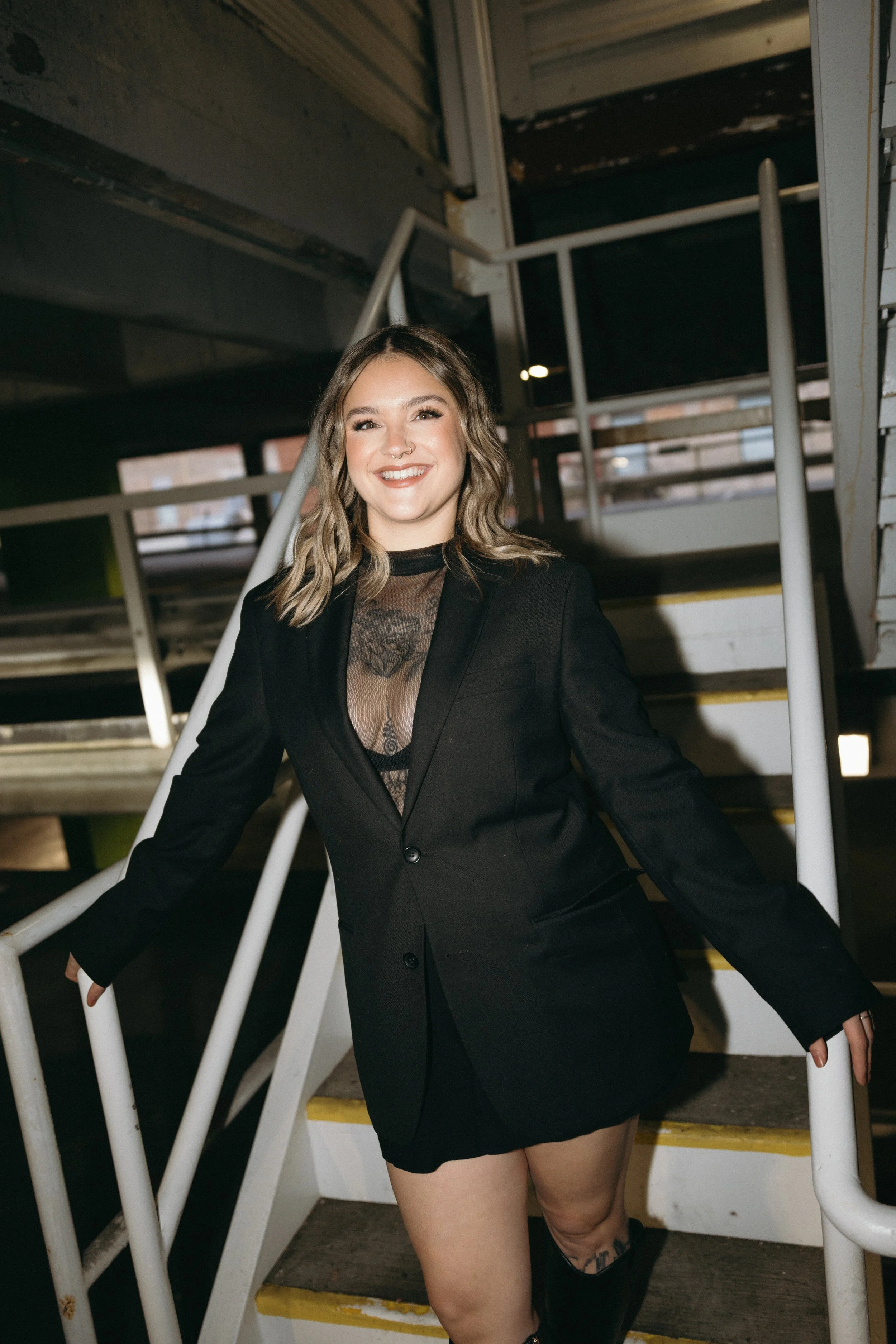A young woman with wavy brown hair smiling at the camera, wearing a black blazer, a sheer black top, and black shorts, standing on a staircase indoors.