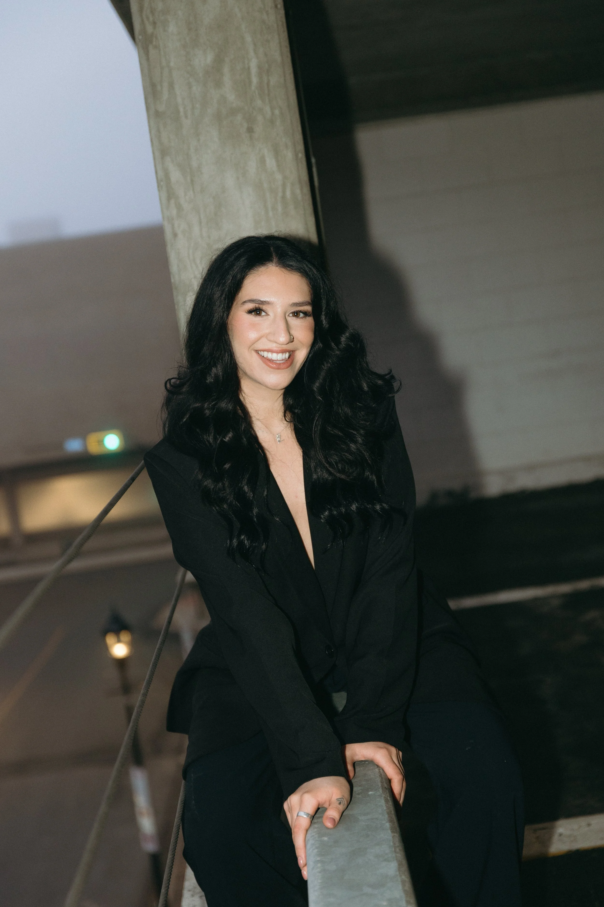 A smiling woman with long, dark, wavy hair is sitting on a railing under an overpass, wearing a black blazer and pants.